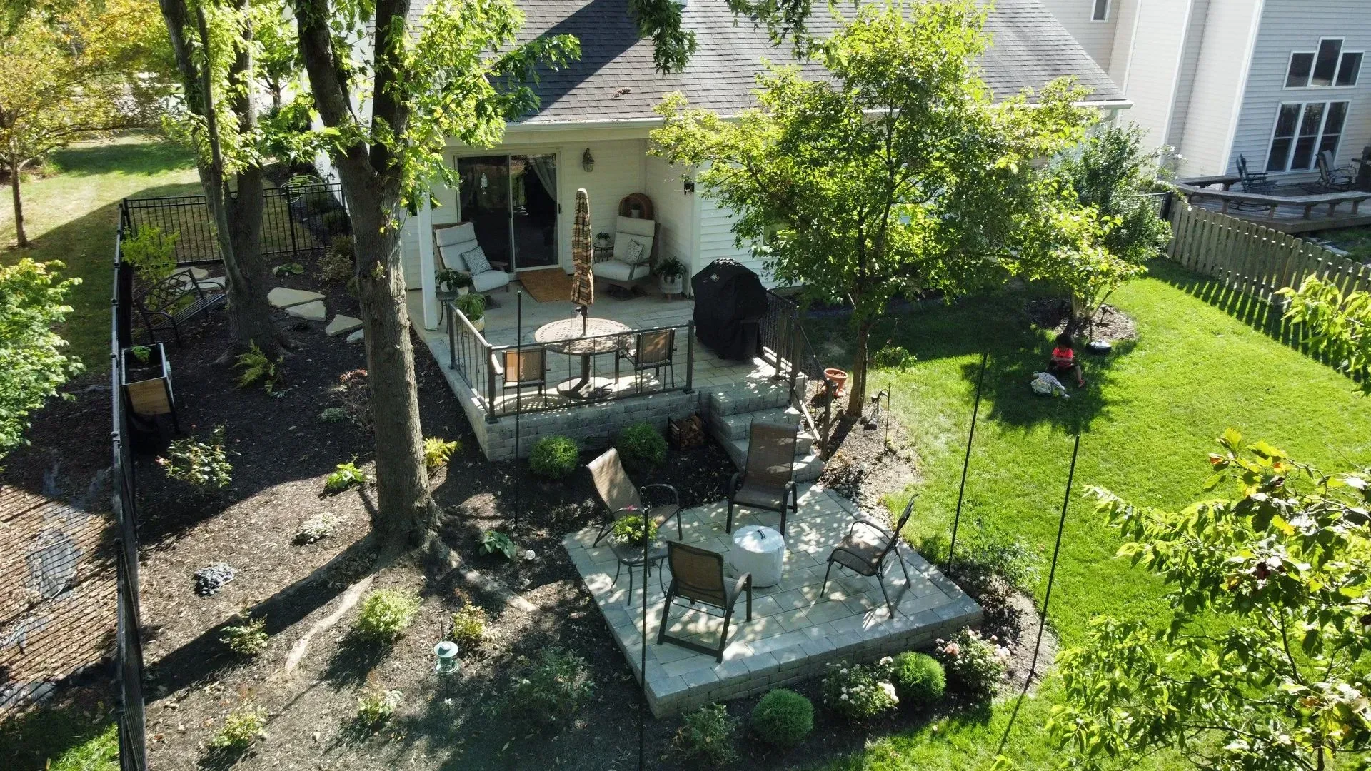 Backyard patio with a table and chairs under a tree beside a white house, surrounded by green lawn and flowers