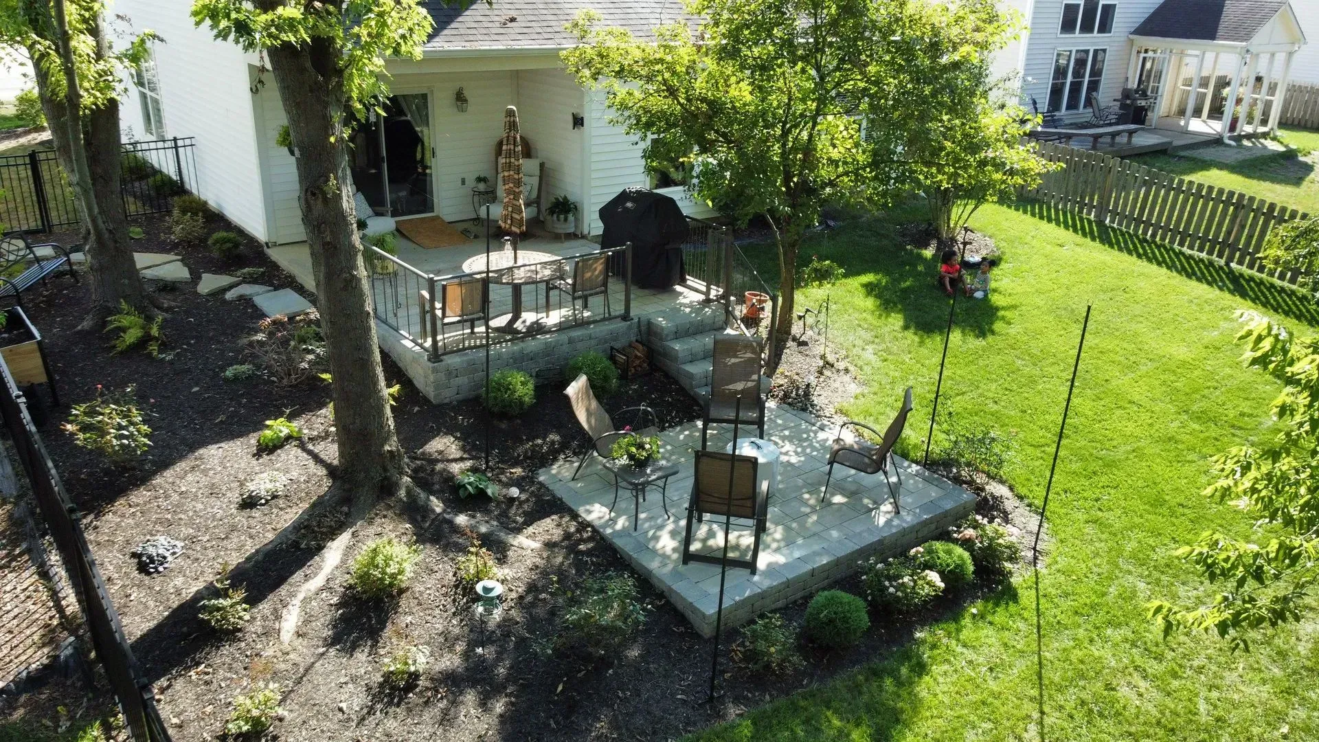 Backyard patio with metal furniture, umbrella, trees, and a green lawn beside a white house