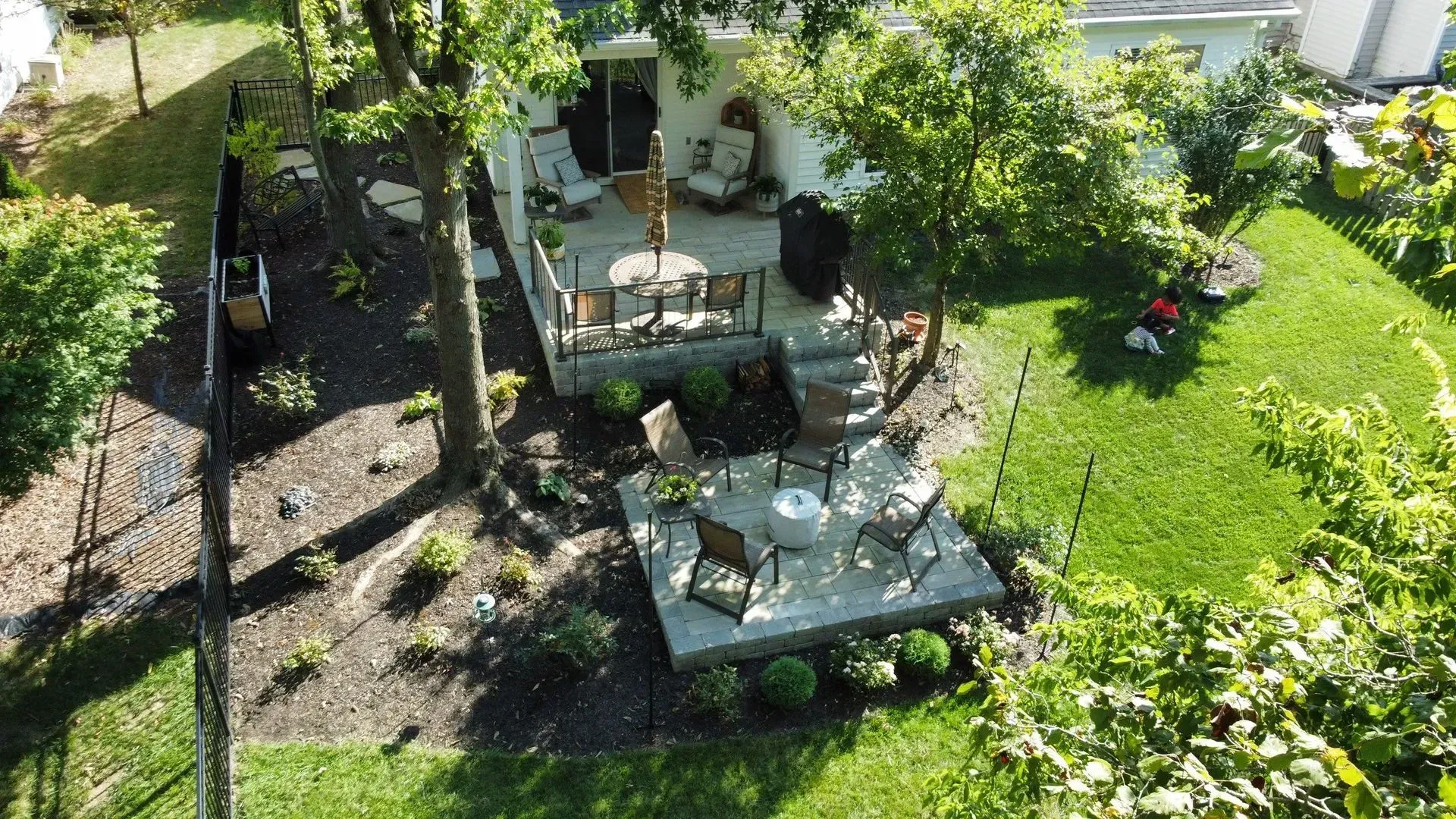 Aerial view of a backyard patio with white tables and chairs, surrounded by green lawn and trees.