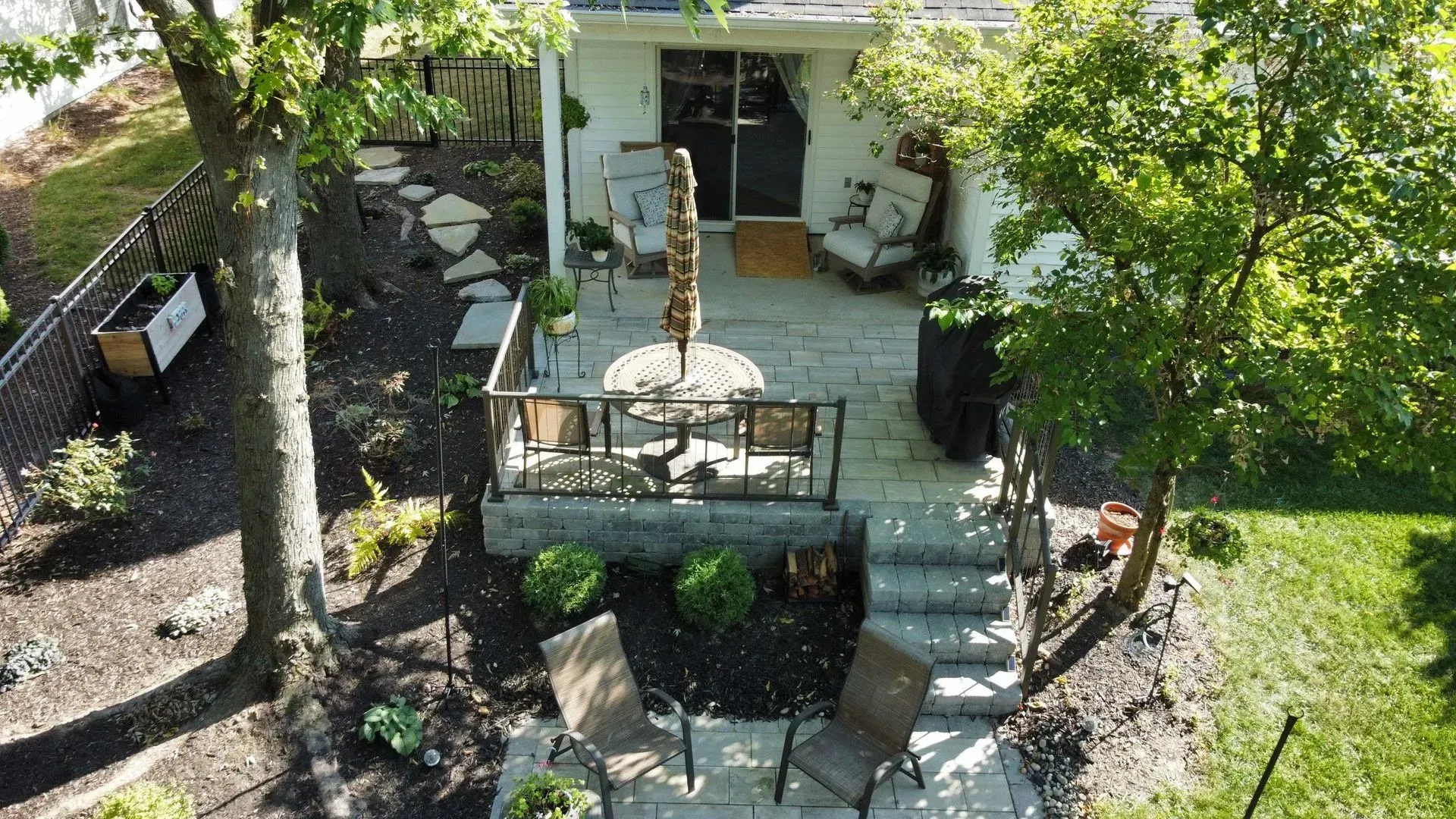 Small patio with table, stairs, potted plants, and surrounding greenery beside a white house