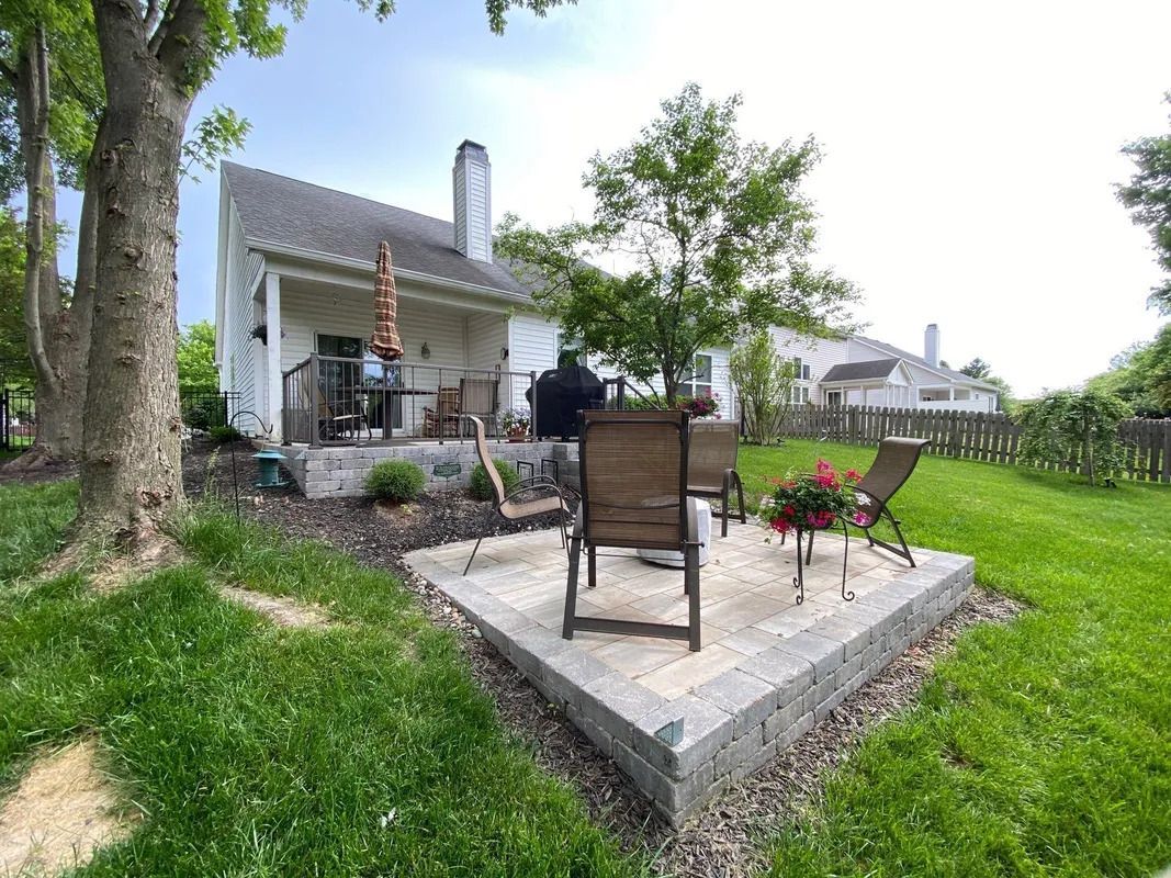Backyard patio with chairs and small table beside a white house, surrounded by green grass and trees
