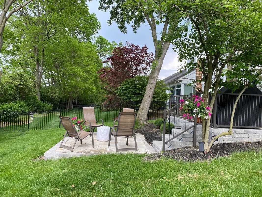 Backyard patio with chairs, table, flowers, and a grill under leafy trees