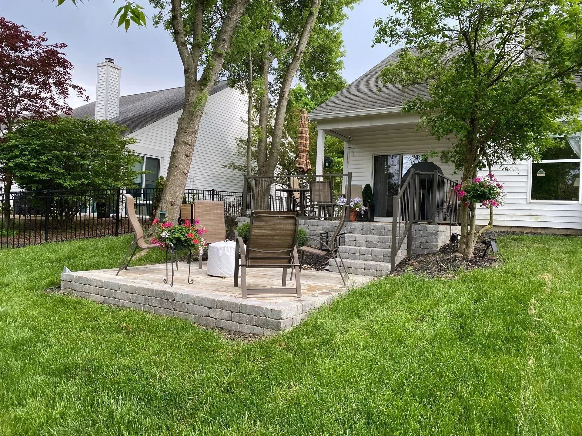 Backyard patio with stone steps, outdoor chairs, flowers, and a white house under trees