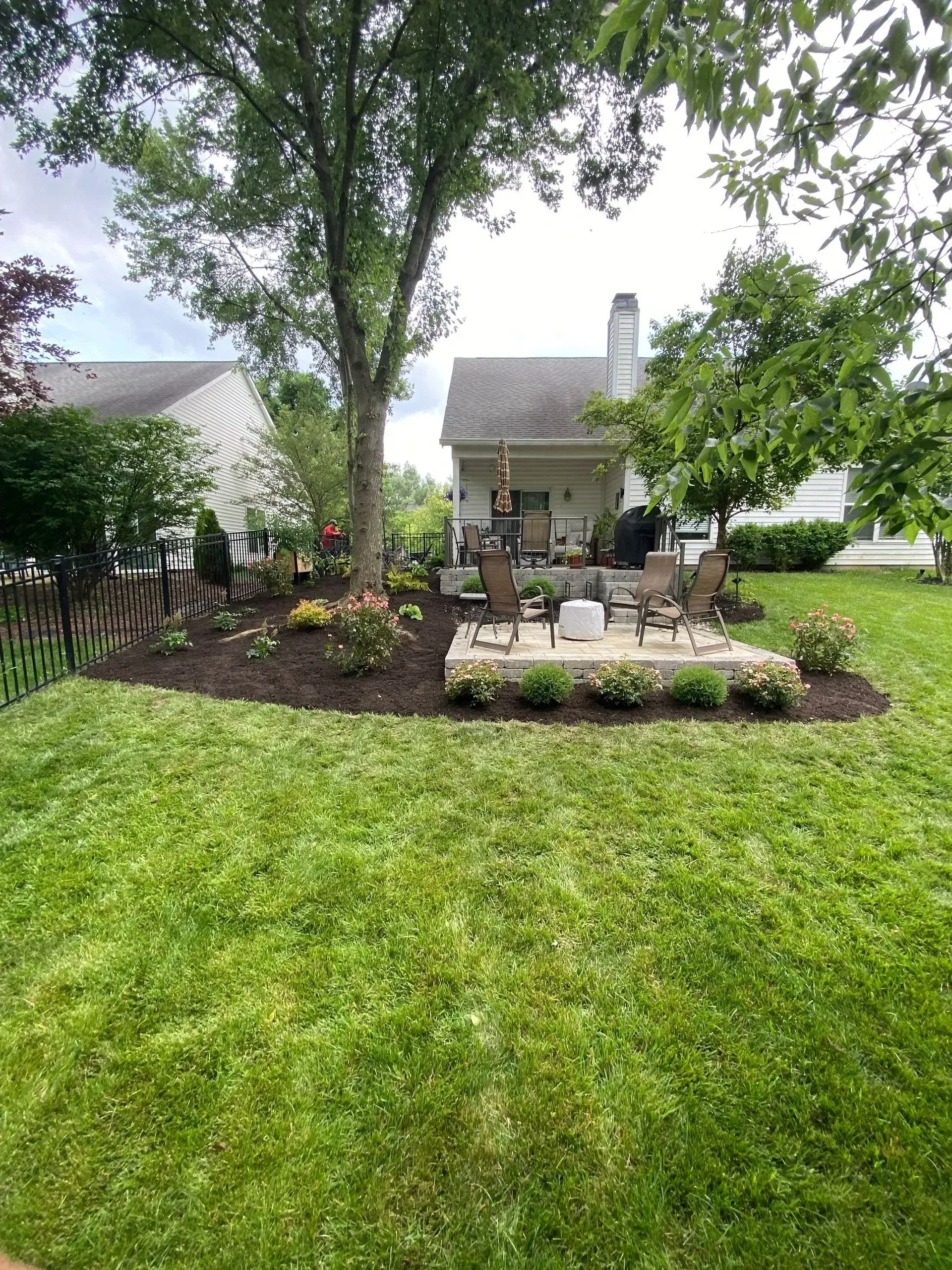 Backyard garden with lawn, flower bed, patio chairs, and a house under cloudy skies