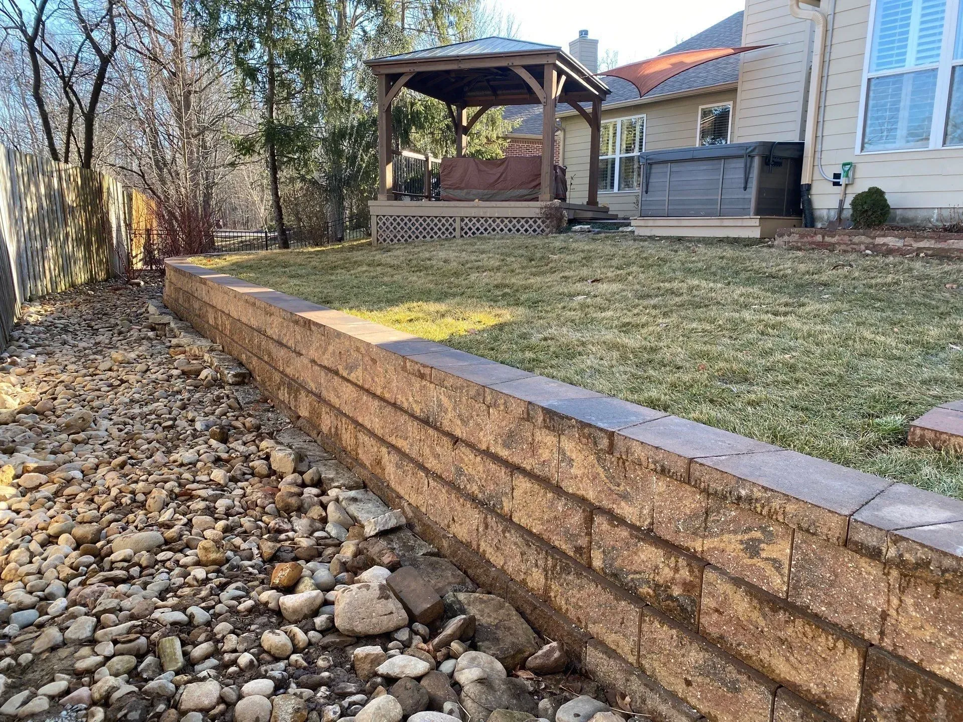 Stone retaining wall bordering a rock bed beside a sloped yard and backyard gazebo.