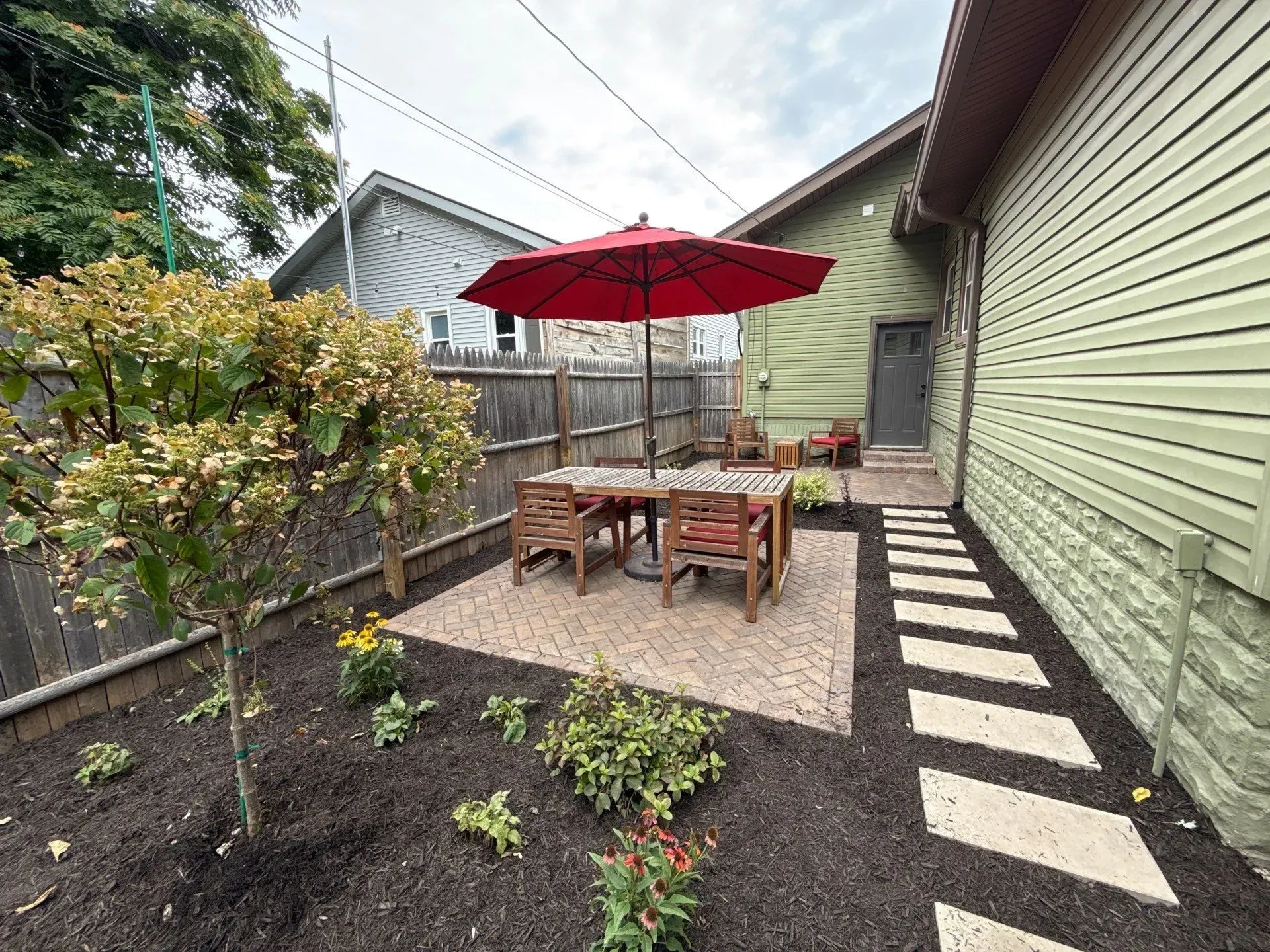 Backyard patio with red umbrella over wooden table, gravel path, and green house exterior.