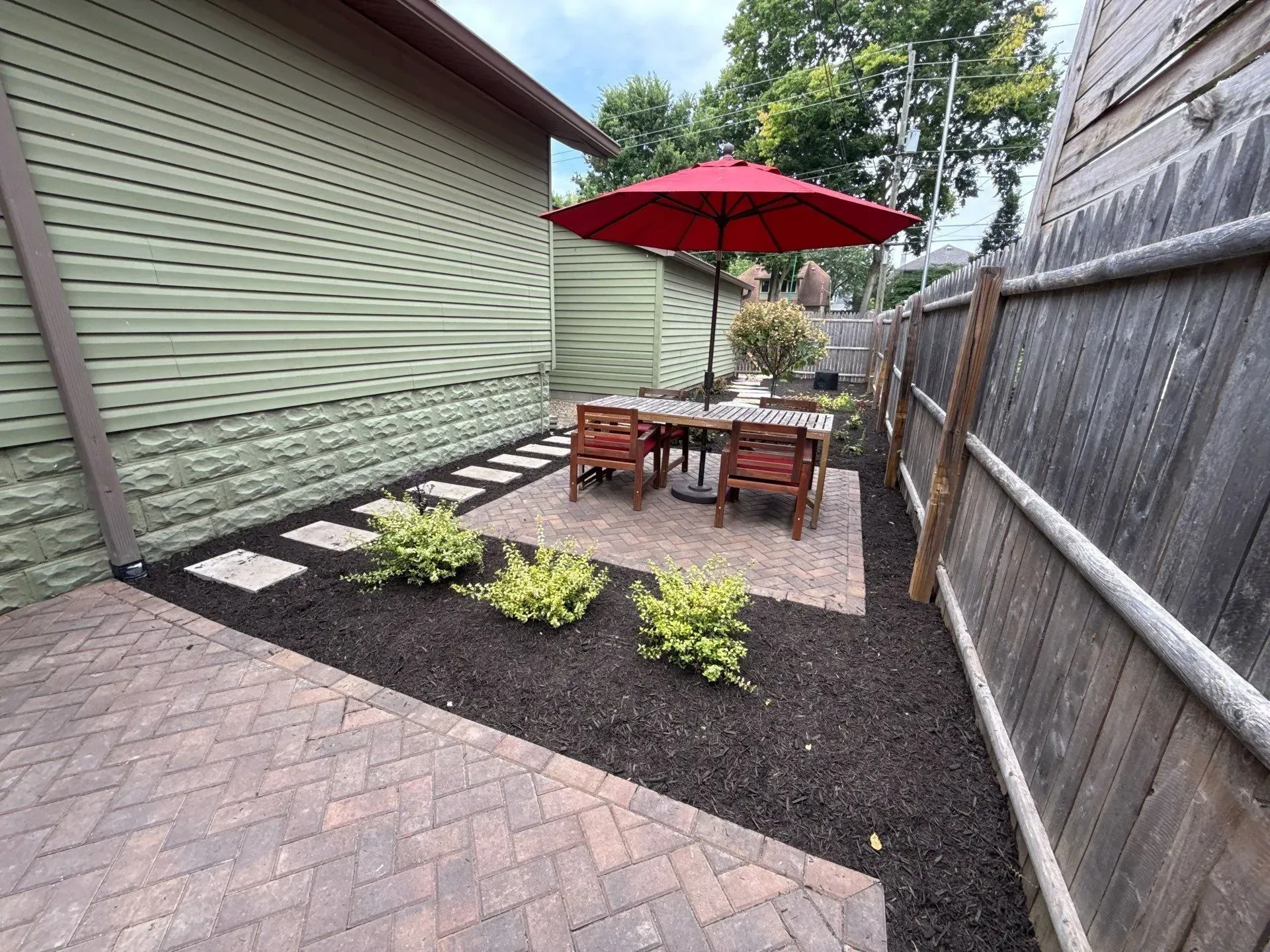 Small backyard patio with red umbrella, outdoor table and chairs, potted plants, and mulch border beside a house and fence.