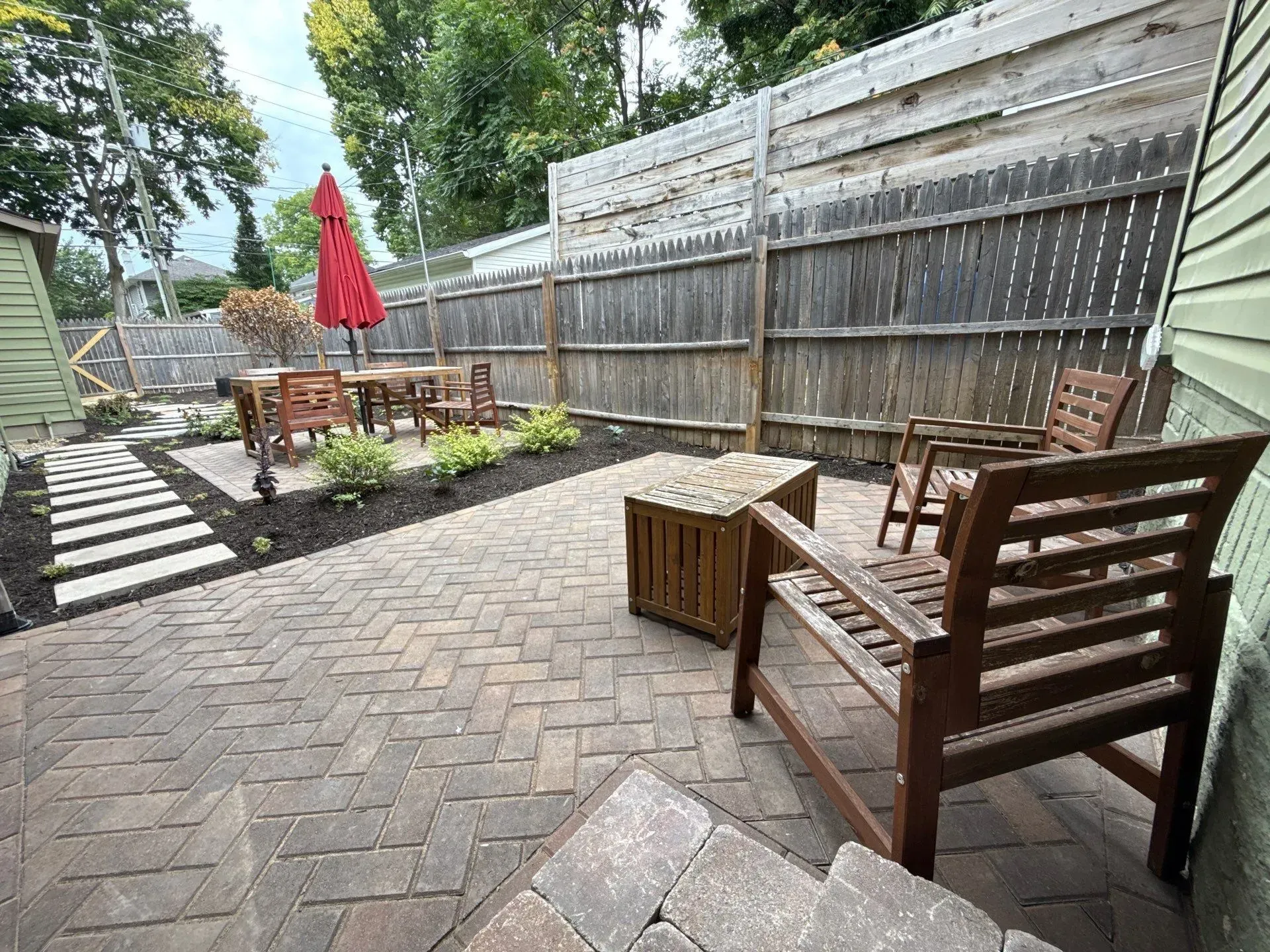 Backyard patio with wooden benches, table, red umbrella, and potted plants beside a fence