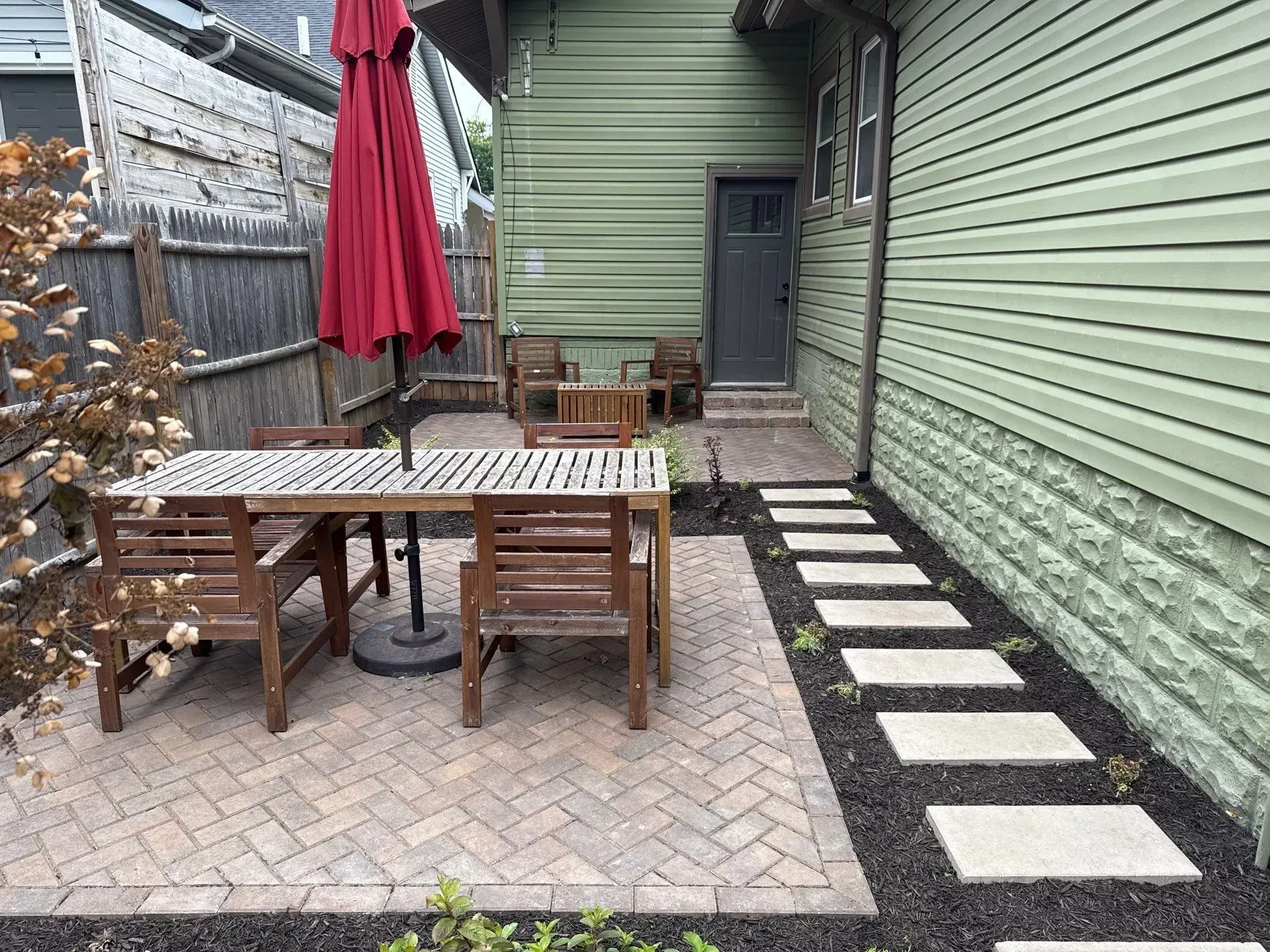 Small backyard patio with wooden table, red umbrella, chairs, and stepping stones beside a green house.