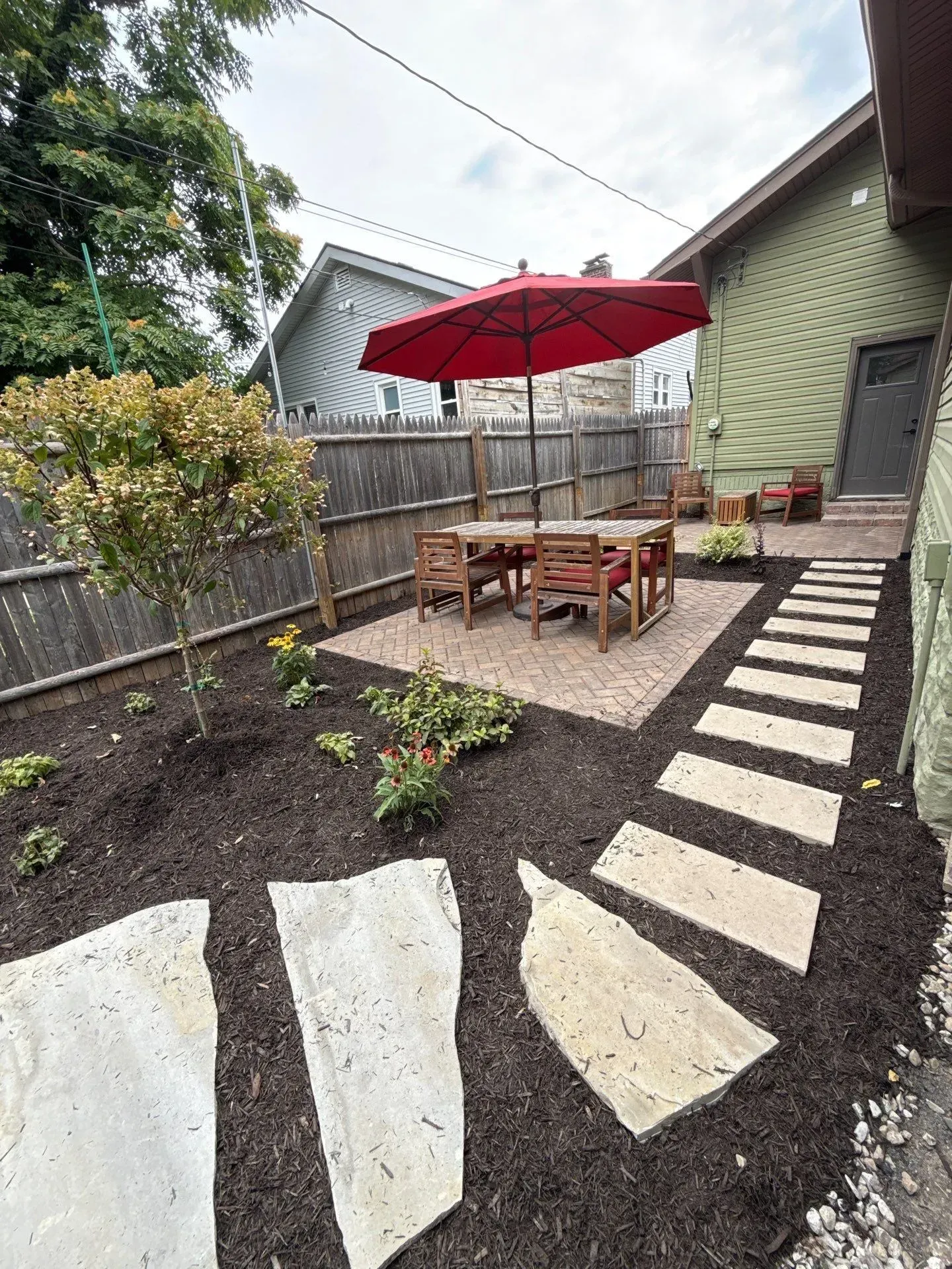 Backyard patio with red umbrella, wooden table, stone path, and stepping stones beside a fence