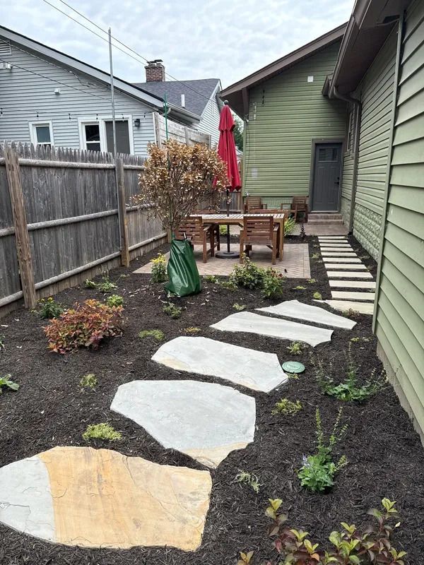 Small backyard garden path with stepping stones, patio table, red umbrella, and wooden fence.
