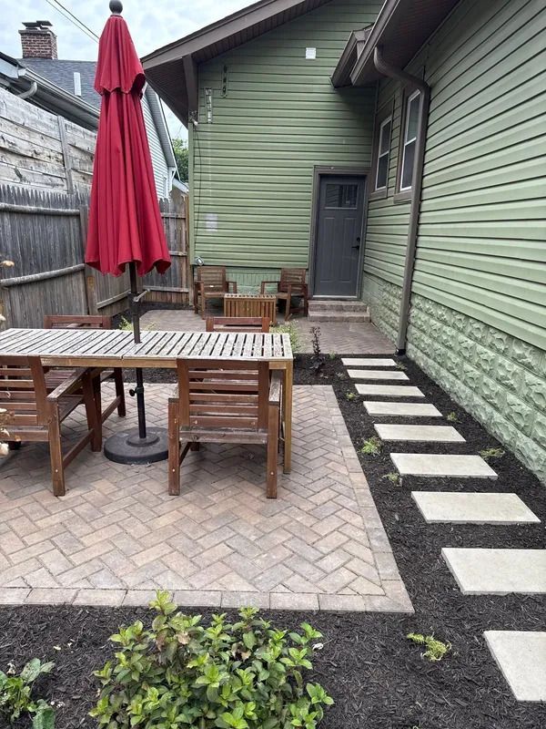 Narrow backyard patio with white stepping stones, dark mulch, red umbrella, wooden table, and fenced sides