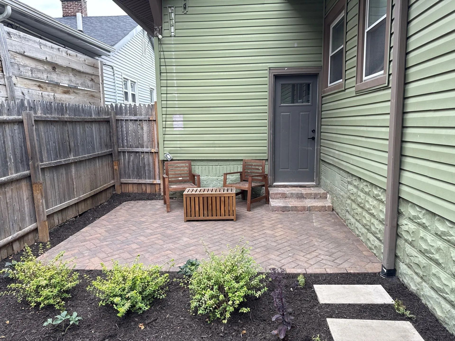Narrow backyard patio with green siding, gray door, wooden chairs, a small table, and gravel ground.