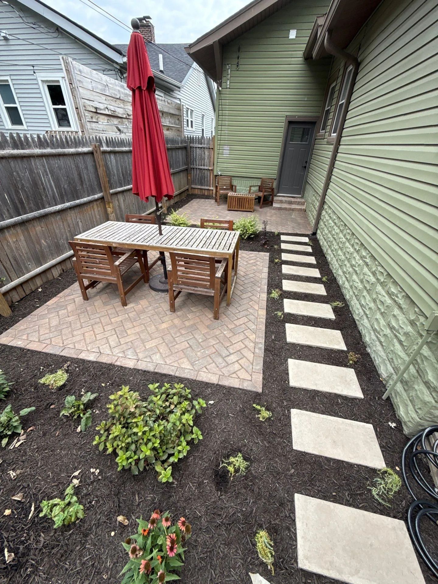 Small backyard patio with dining table, red umbrella, potted plants, and stepping stones along a green house.