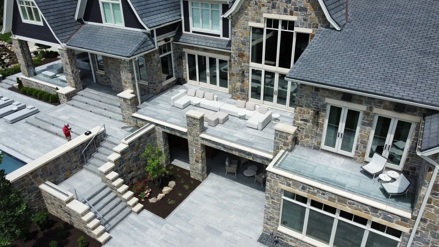 Aerial view of a large gray stone house with patio, stairs, and a person in red on the left terrace
