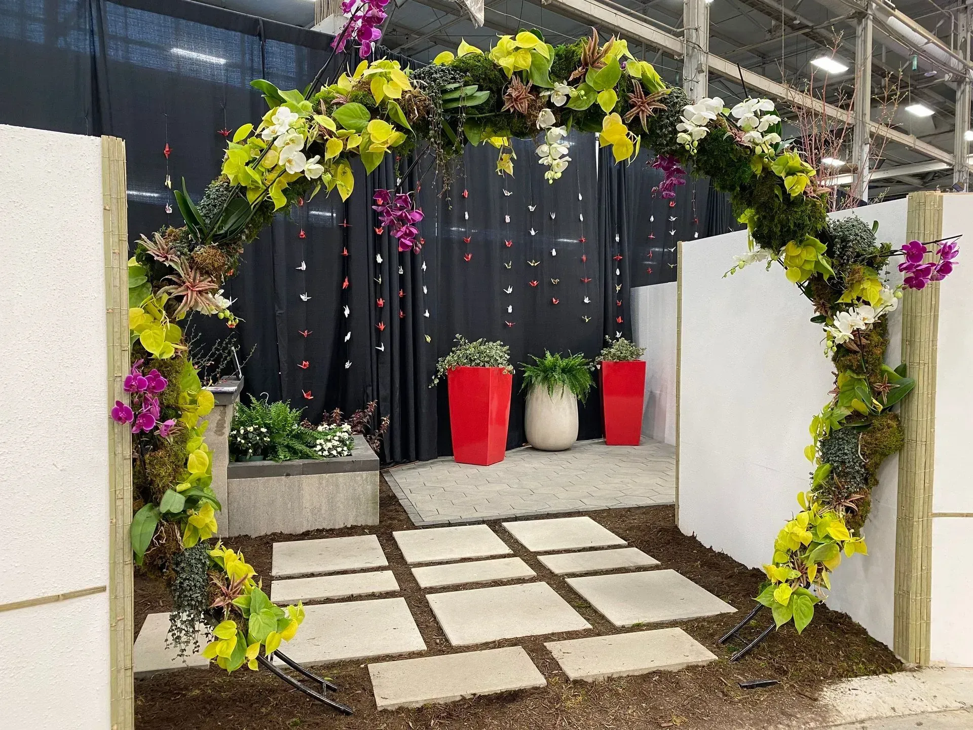 Flower-covered archway with white stepping stones leading to a display area with red and white planters