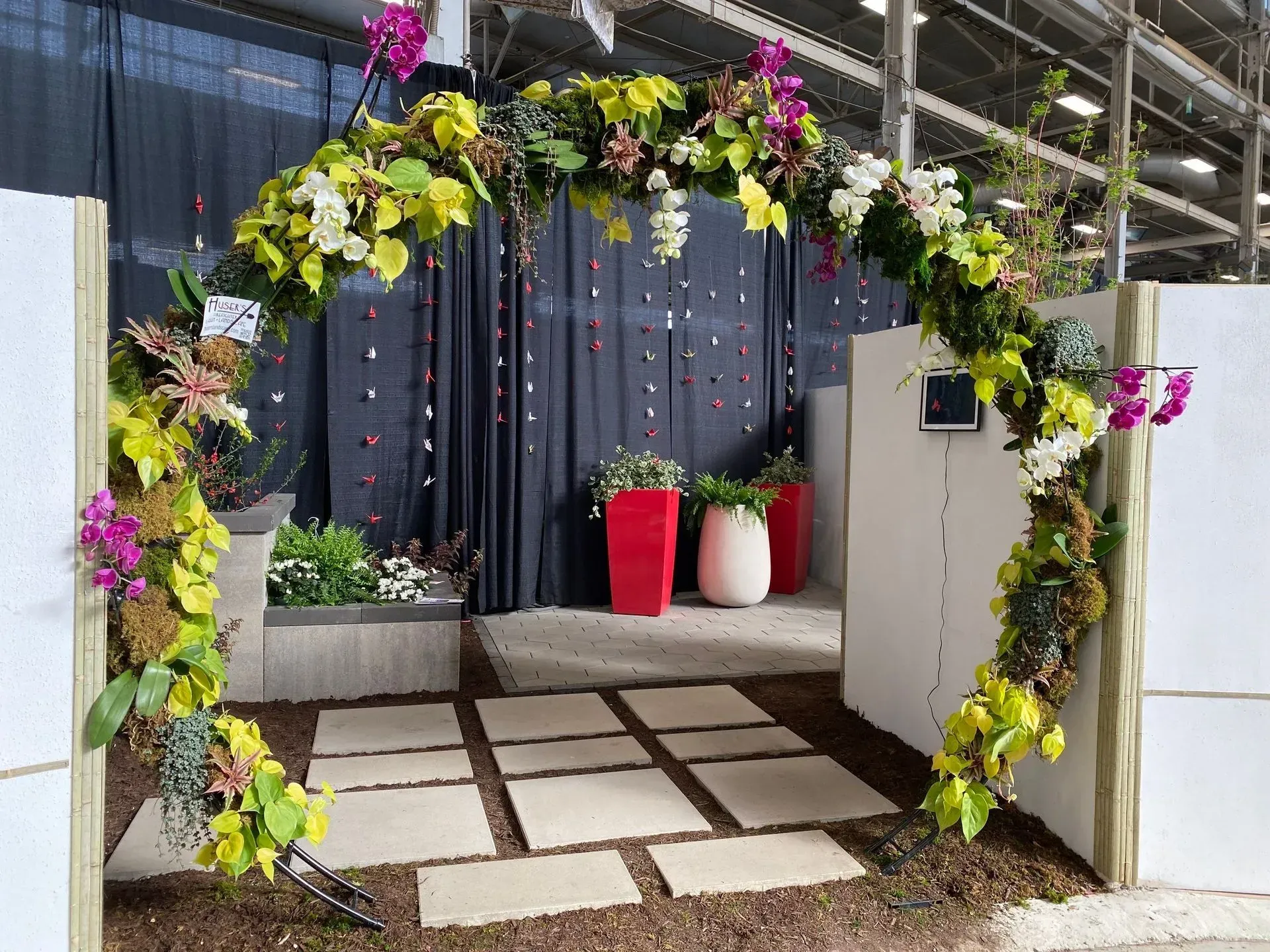 Flower-covered archway framing a patio entrance with white pavers, hanging plants, and red and white planters