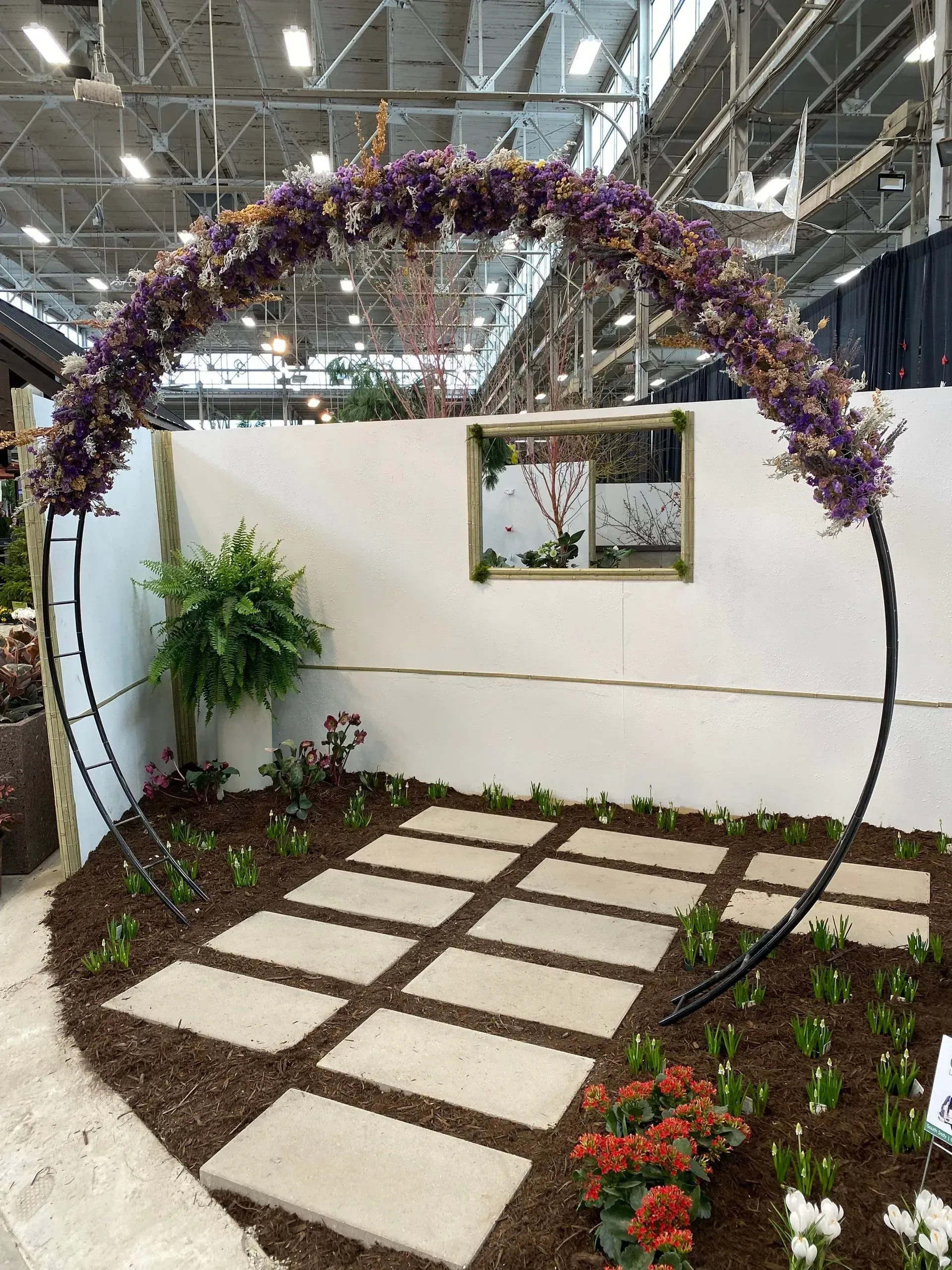 Decorative garden display with stone pavers, plants, and a purple floral arch in an indoor expo hall
