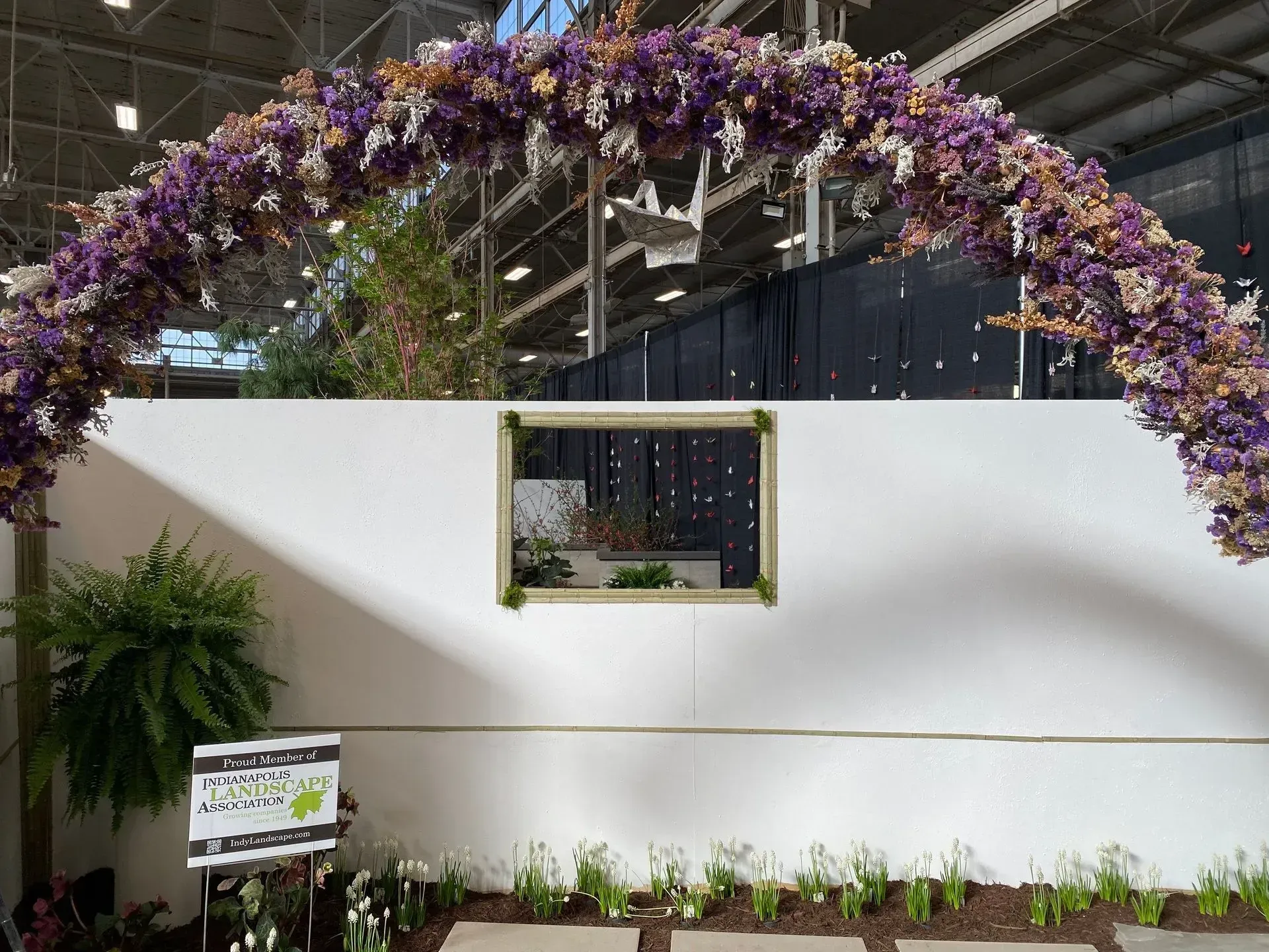 Purple flower arch over a white garden wall with a small window and potted plants below