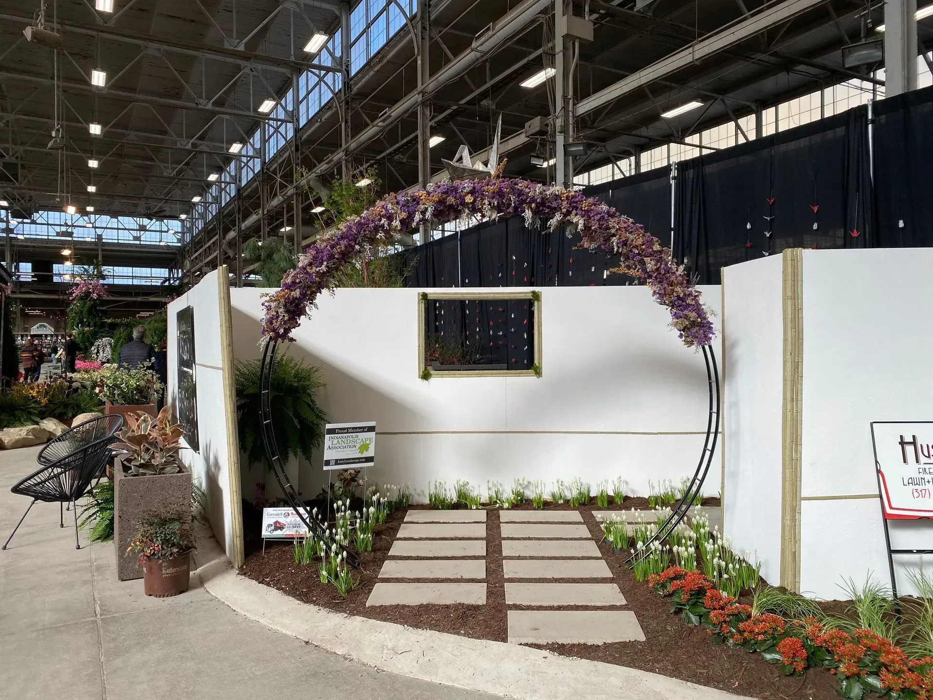 Flower-covered arch over a small garden display in an indoor hall with a white wall and tiled path.