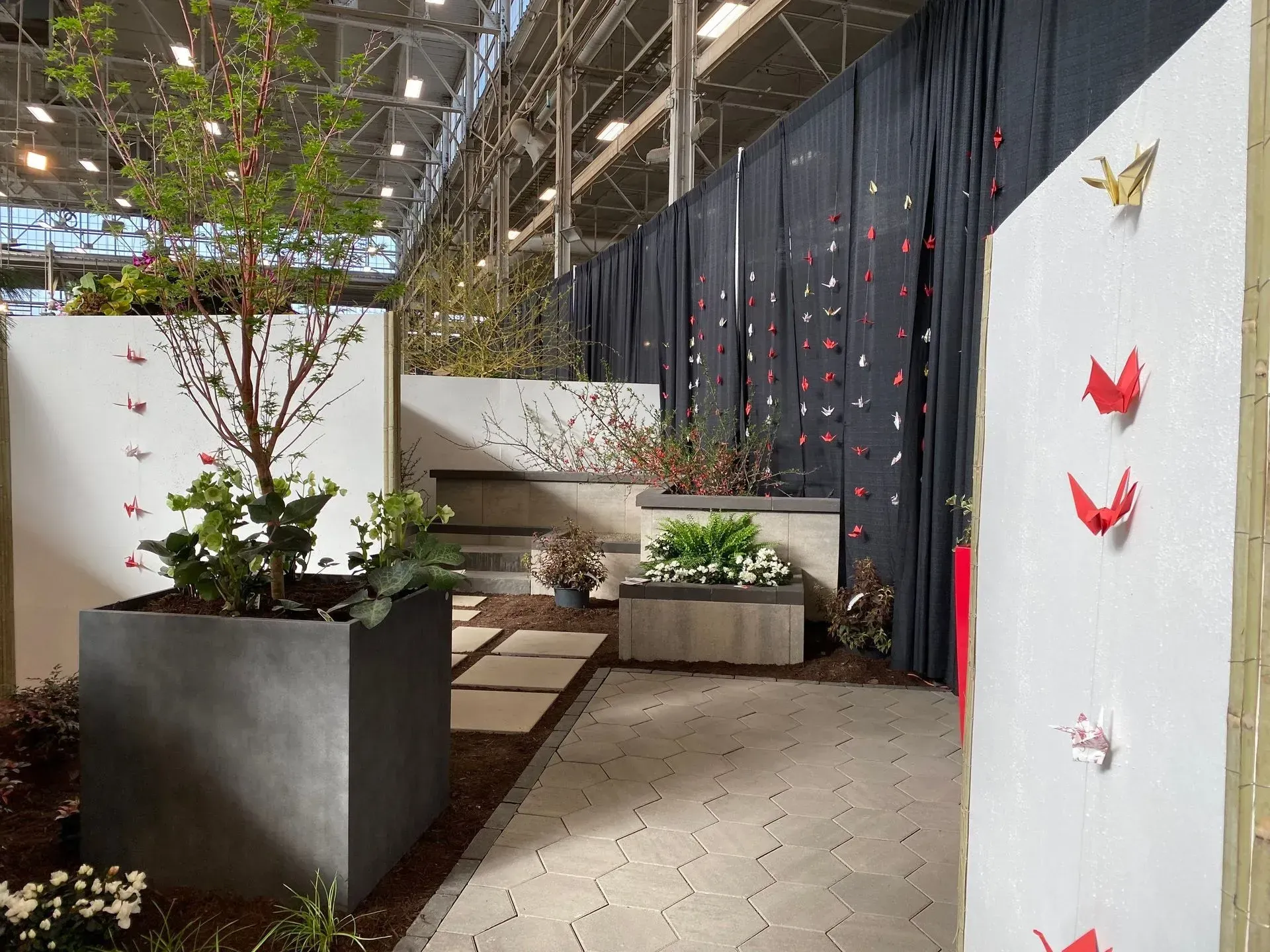 Indoor exhibition space with potted trees, white panels, and red origami birds on a wall