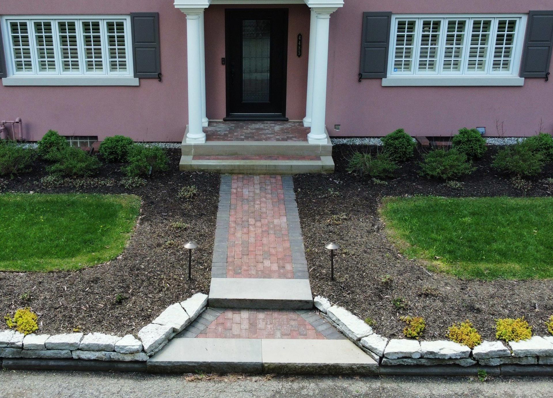 Brick walkway leads to a pink house with white columns, windows, and landscaping.