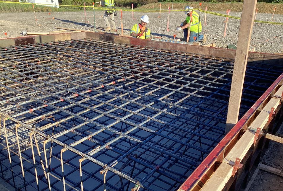 Construction workers inspecting rebar grid within concrete formwork. Outdoors, sunny.