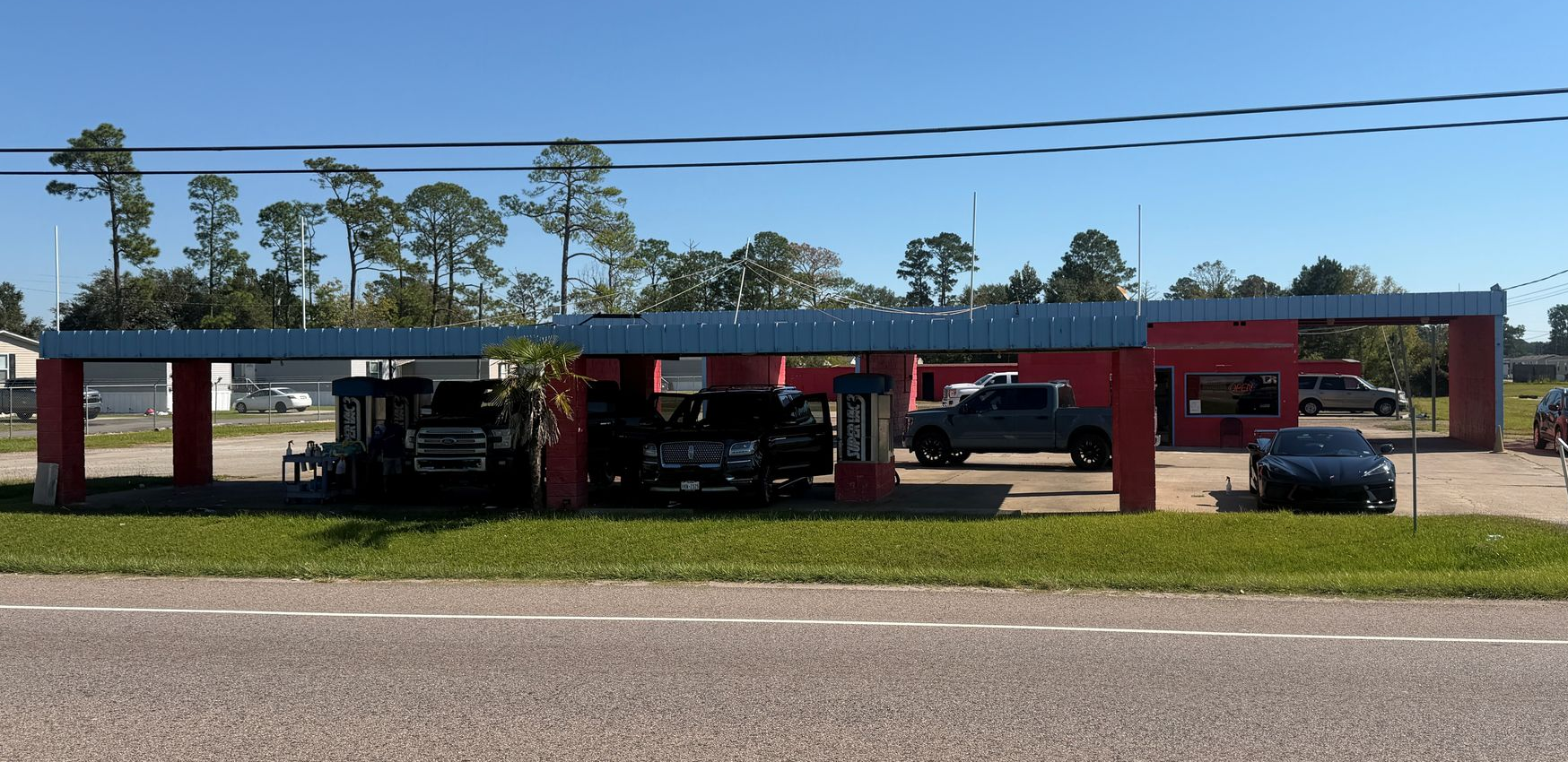 Drive-through car wash with red pillars, blue roof, and vehicles in bays under a clear, sunny sky.
