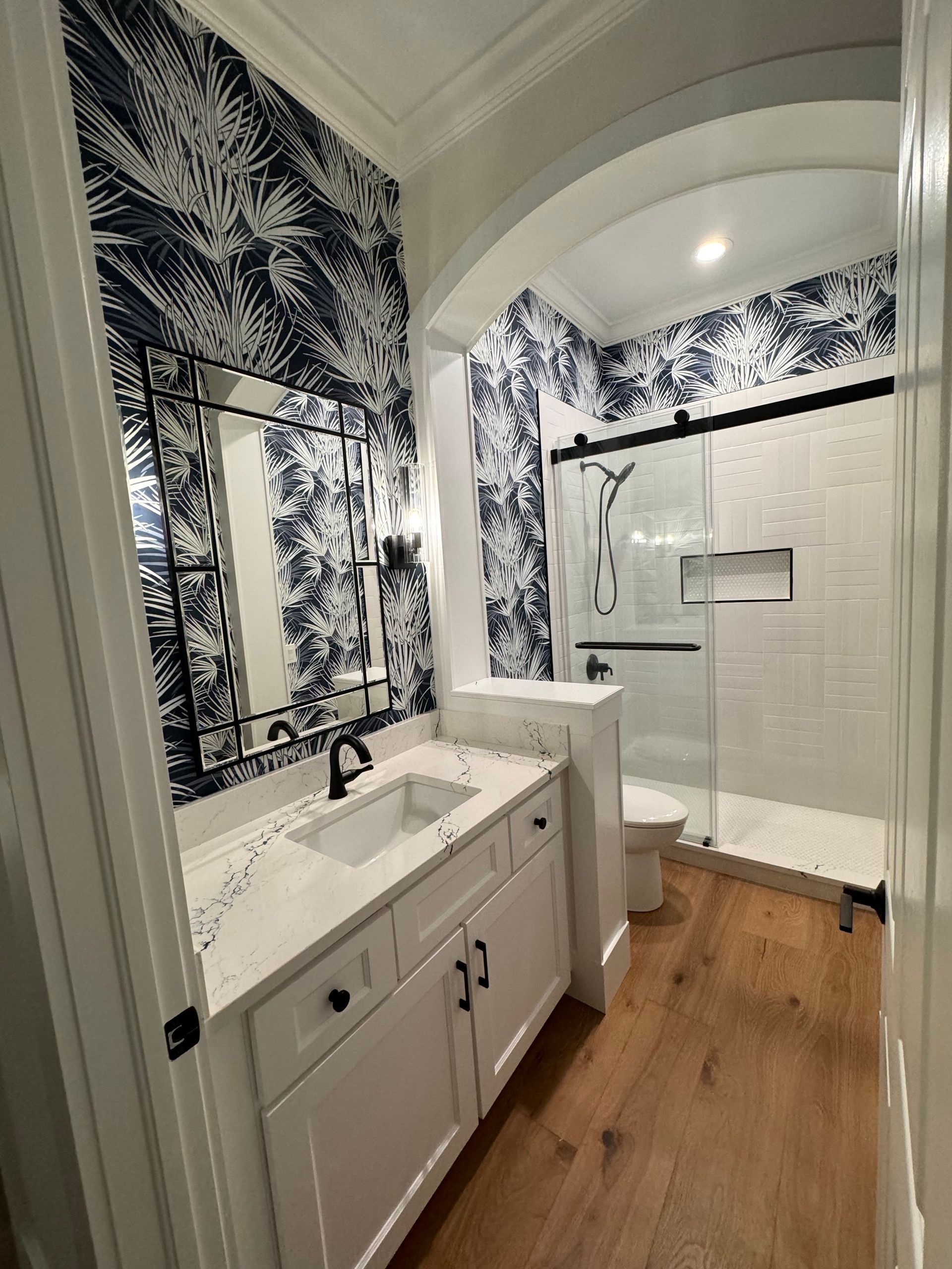 Bathroom with white vanity, patterned blue wallpaper, arched entry to a shower, and wood flooring.