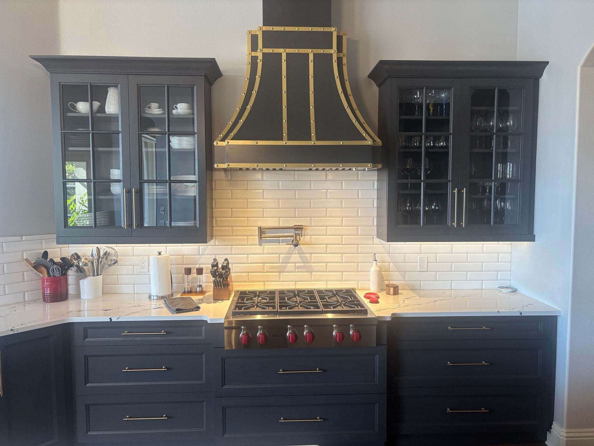 Kitchen with dark cabinets, a stove, a black and gold range hood, and white tile backsplash.