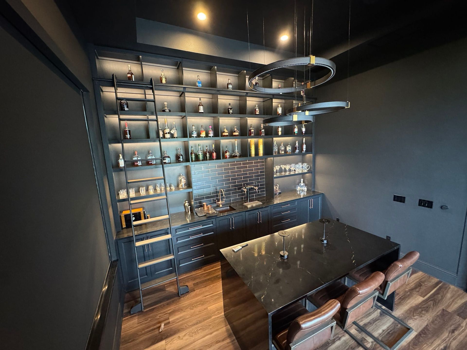 Dark bar area with a large liquor cabinet, counter, table with seating, and modern lighting.