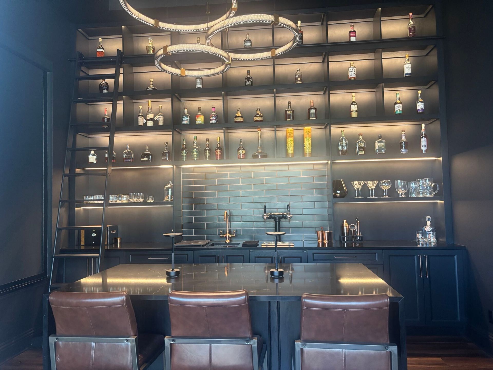 Home bar with black shelving, lit bottles, black countertop, brown leather chairs, and gold fixtures.