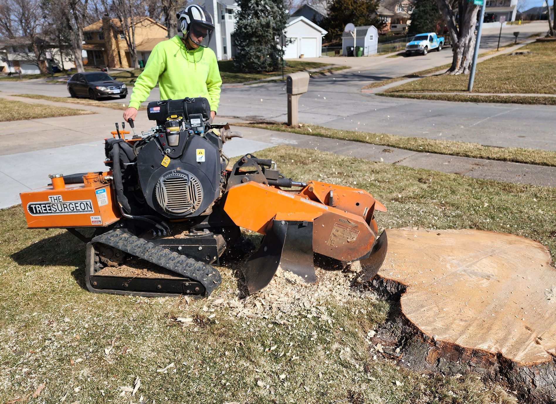 Man operating stump grinder on lawn, residential street in background.