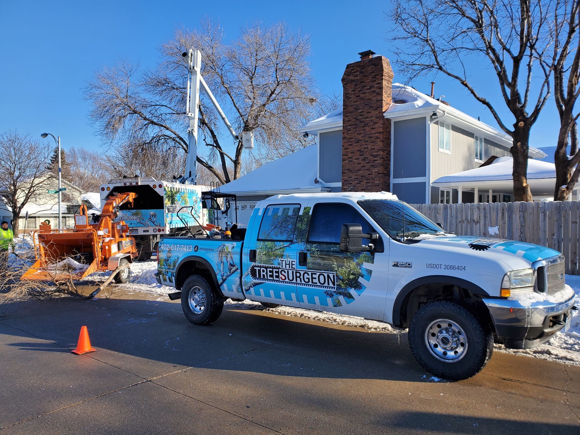 Tree service truck with wood chipper on a snowy street, removing branches near a house.