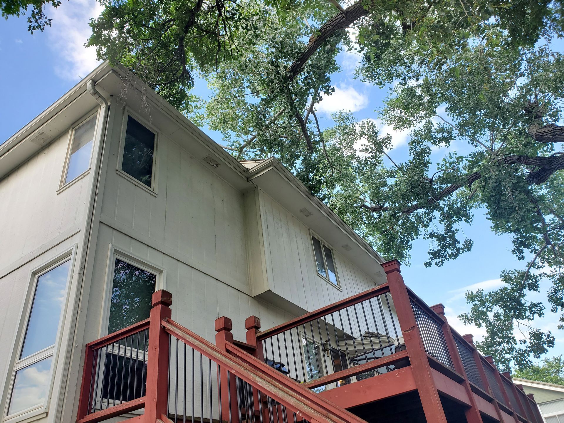 Beige two-story house with a wooden deck, black railings, and tree branches against a blue sky.