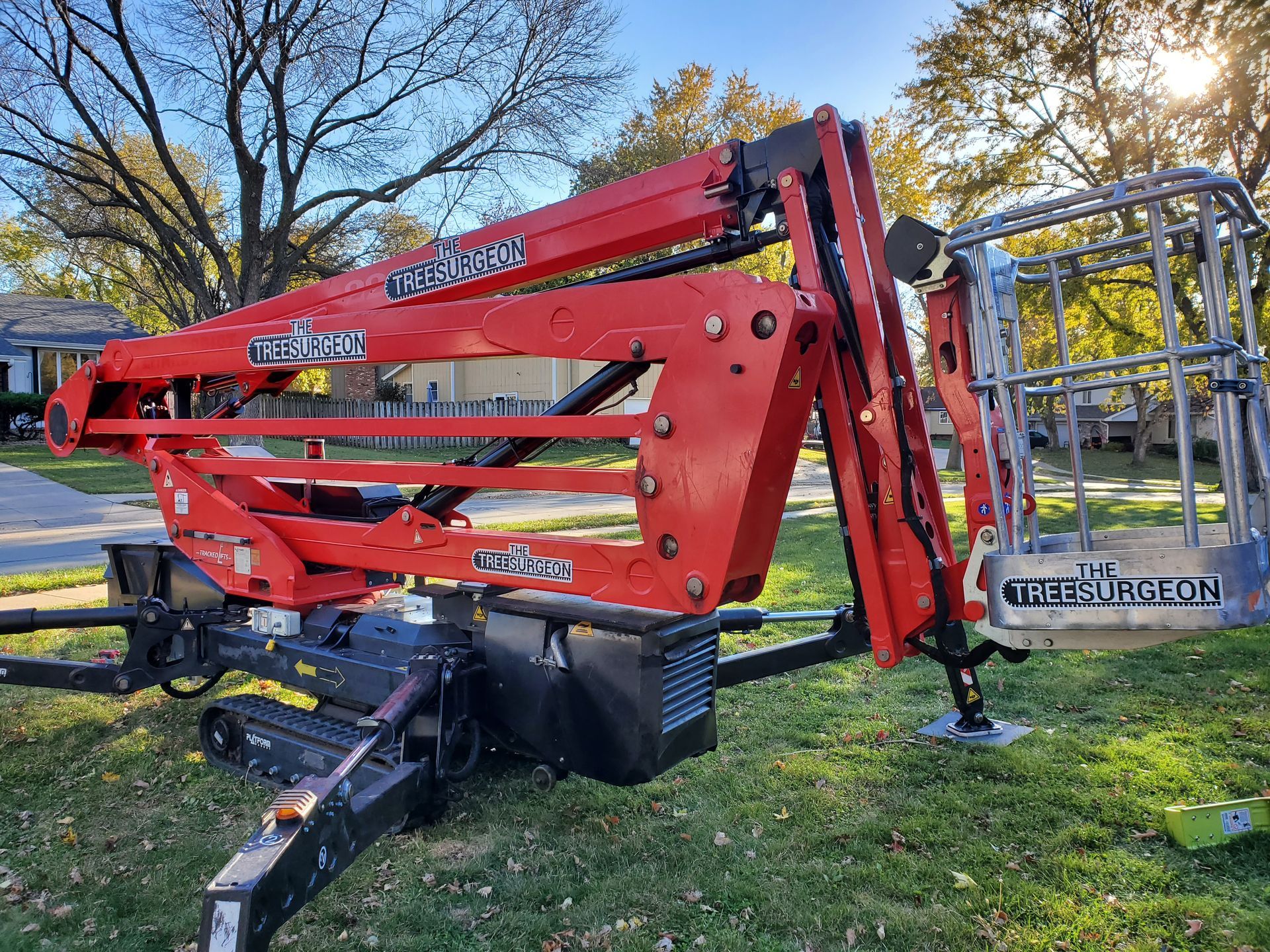 Red towable boom lift on grass, with an empty work basket extended; trees in the background.