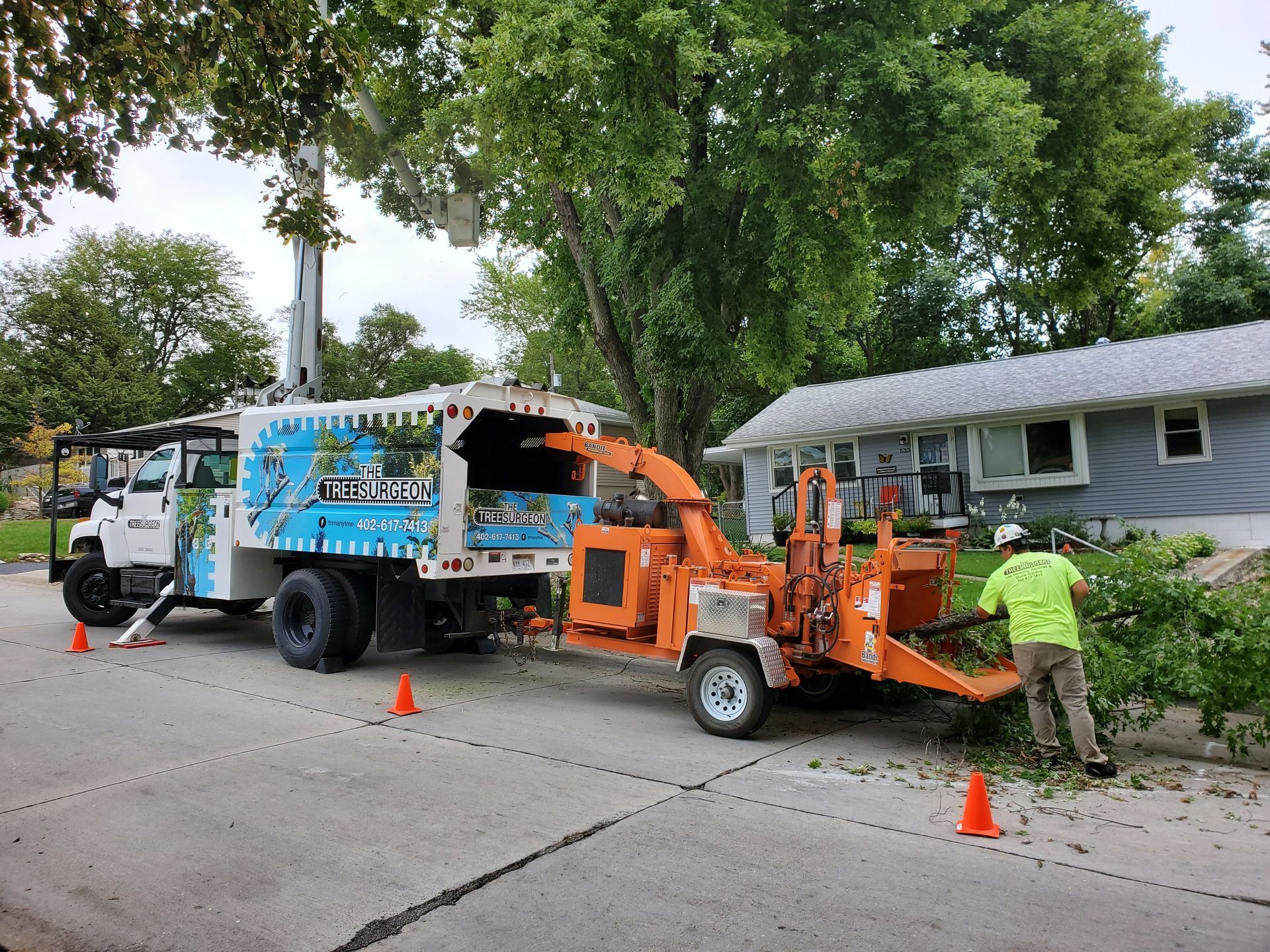 Arborist chipping branches on a residential street. Orange chipper, tree trimming truck, worker in neon green shirt.