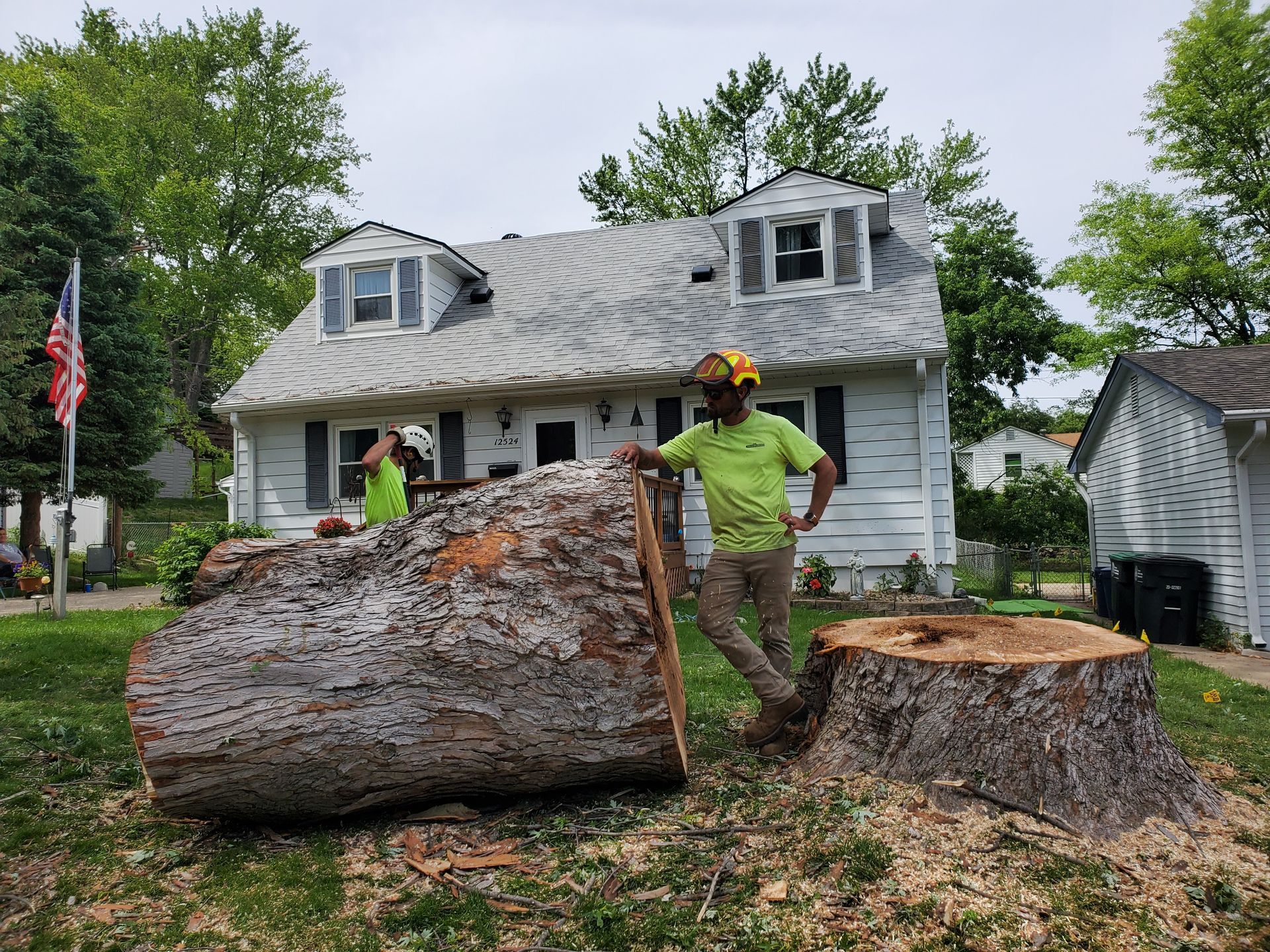Two people near a fallen tree trunk and stump in front of a house. One person wears a helmet.