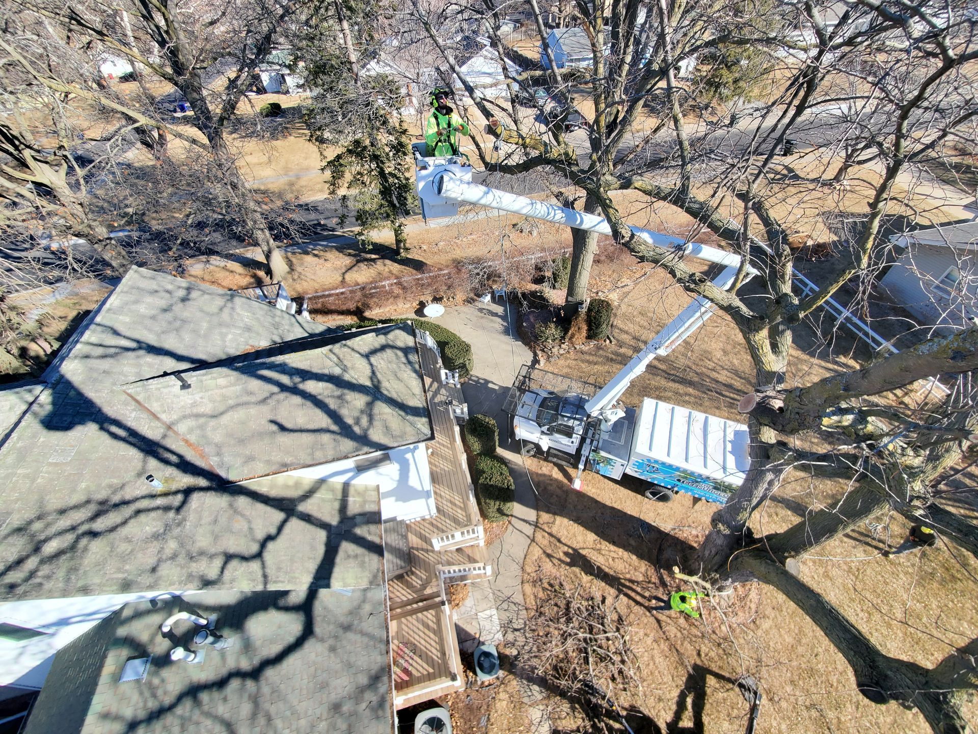 An aerial view shows a tree being trimmed by a lift truck. The roof of a house is visible in the foreground.