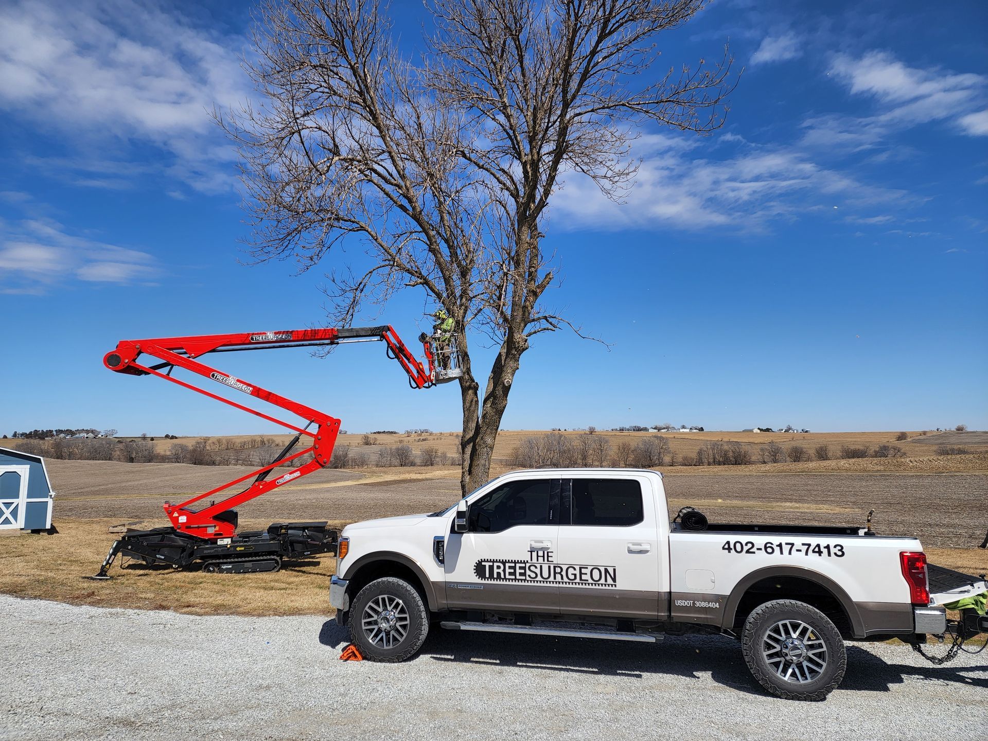 Tree trimming with a red lift truck and white pickup truck under a clear blue sky.