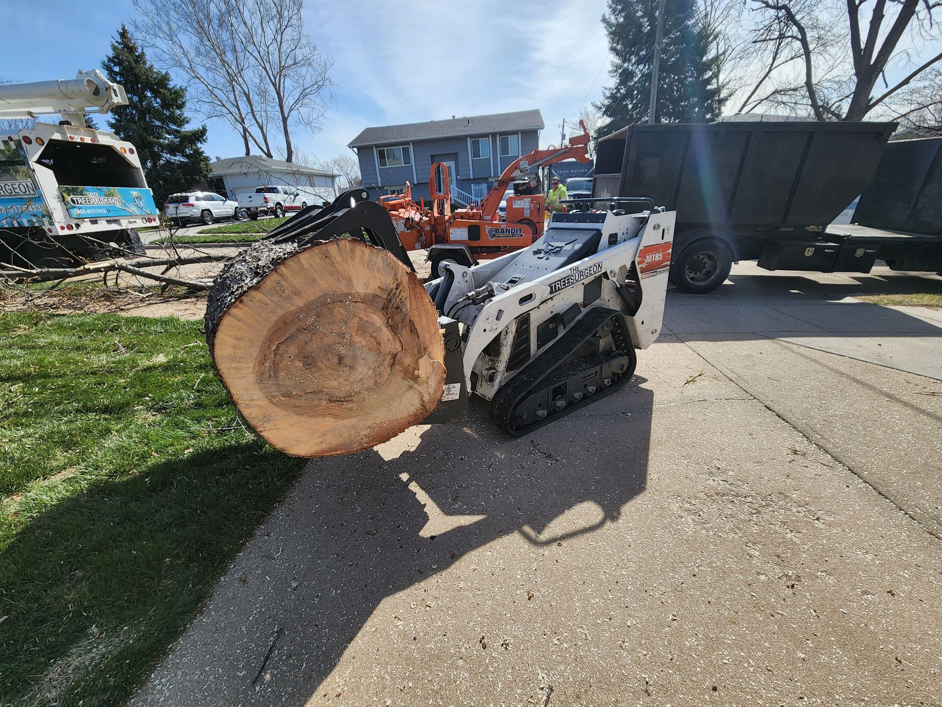 Bobcat skid steer with a large tree trunk on pavement near a chipper and truck, sunny day.