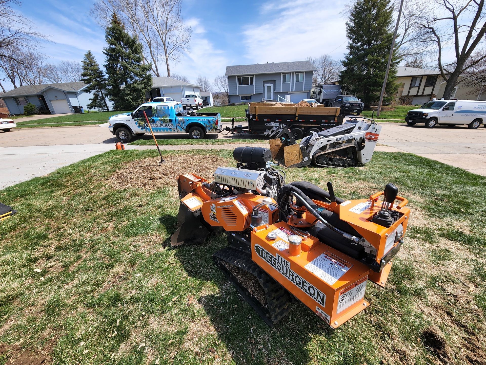 Stump grinder on grass with equipment, truck, and houses in the background on a sunny day.