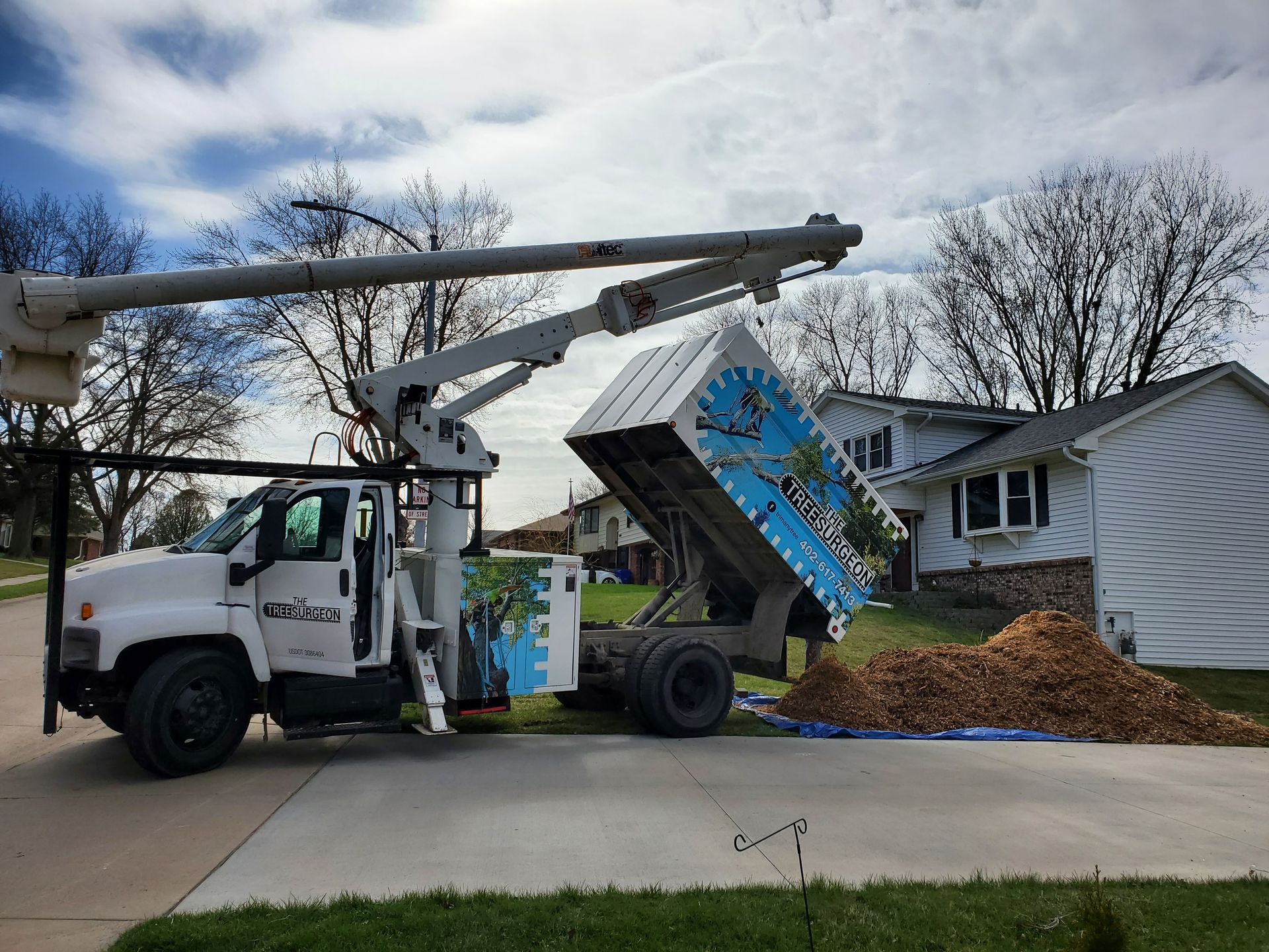 White tree service truck dumping wood chips in a residential driveway, boom extended.