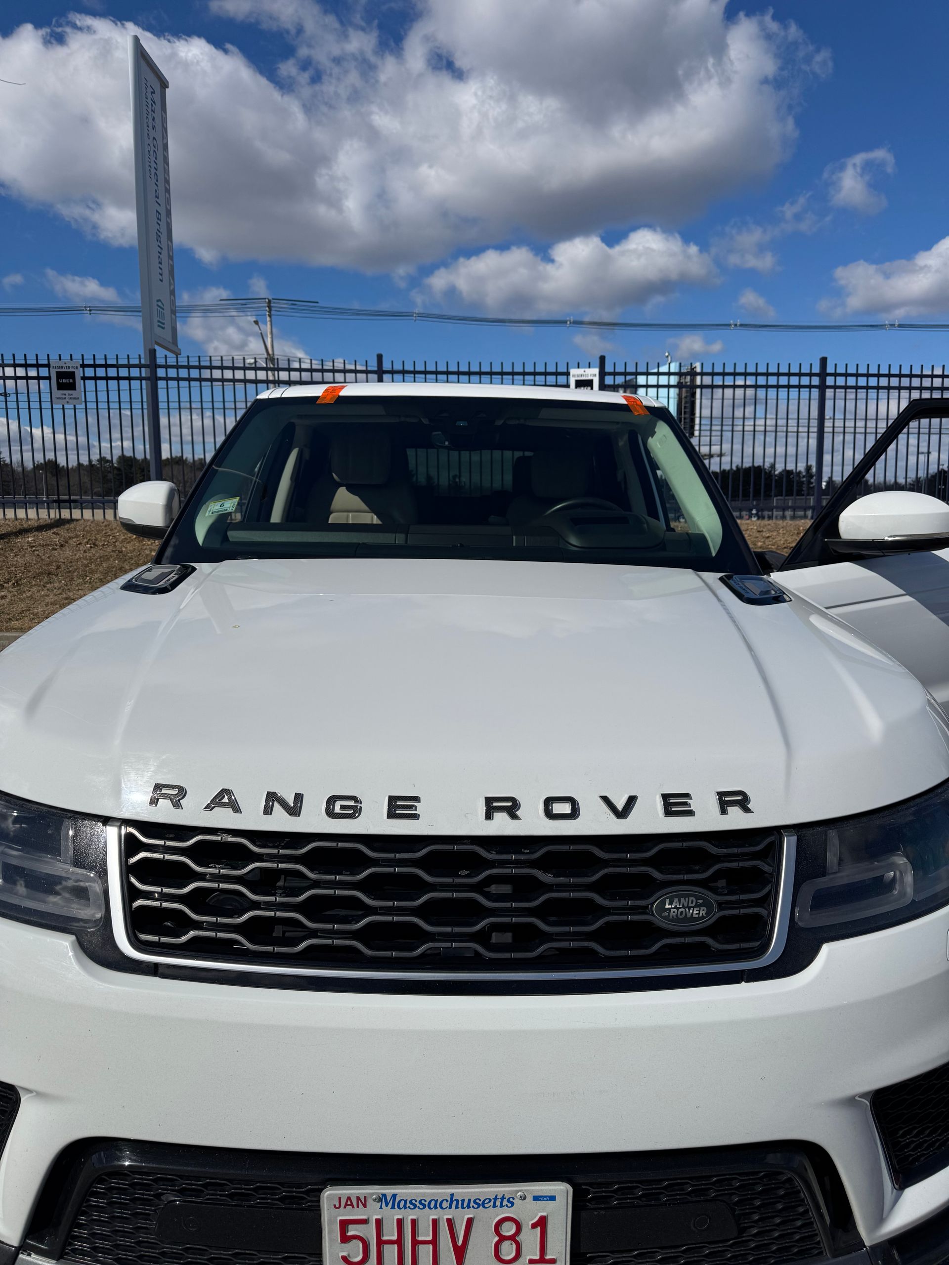 White Range Rover SUV with Massachusetts license plate, parked in front of a fence, under a blue sky with clouds.