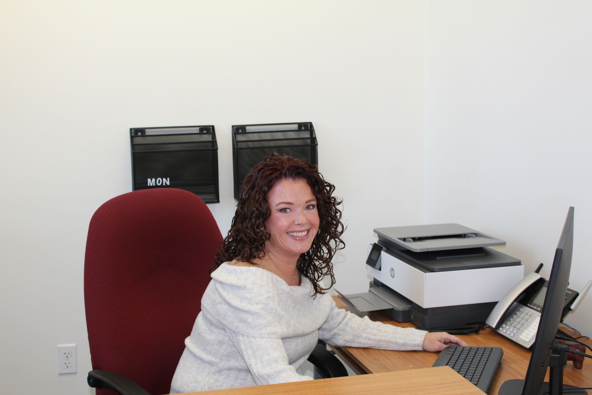 Woman smiling at a desk in an office setting. She is wearing a light sweater and has dark curly hair.