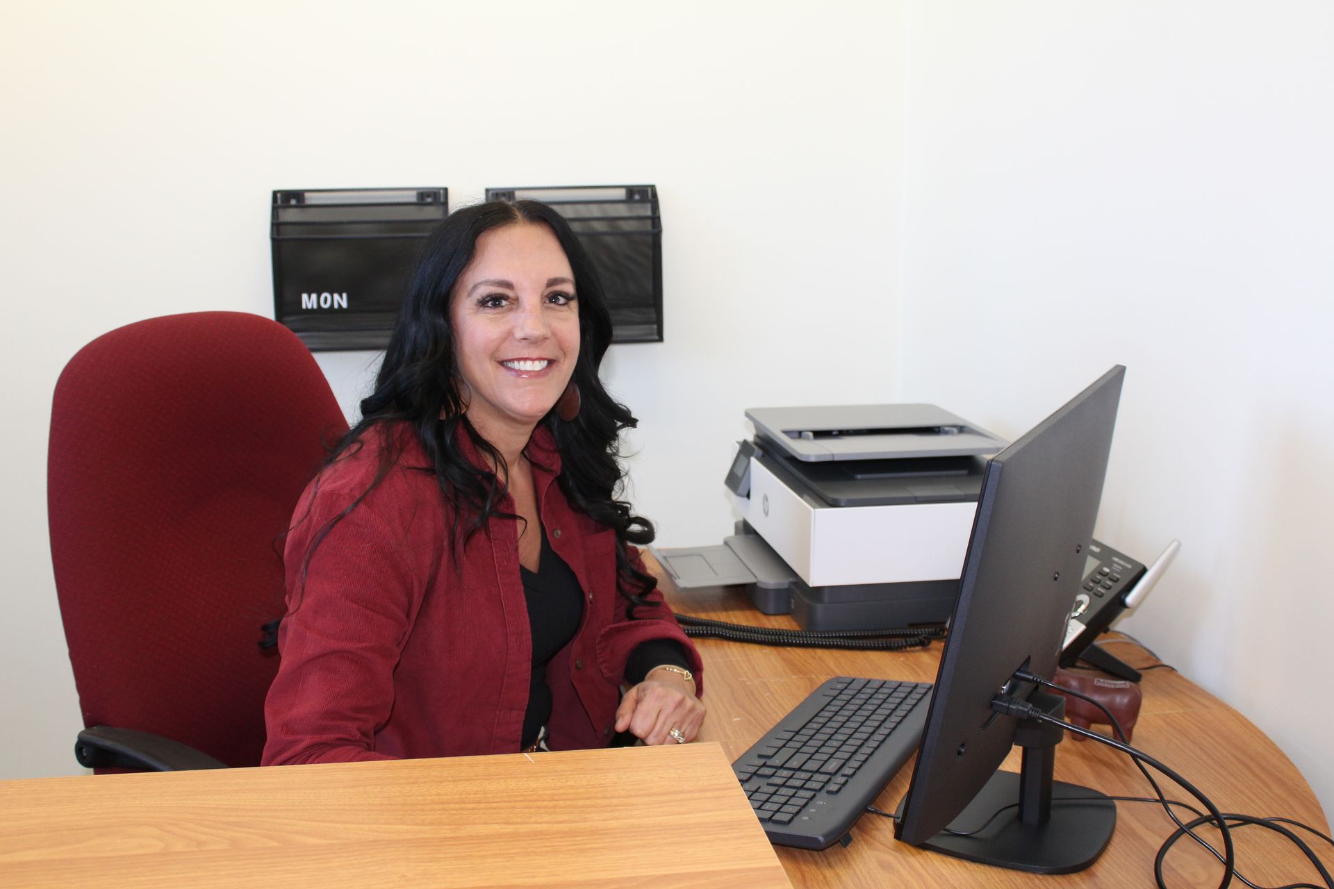 Woman smiles at a desk with a computer, printer, and phone in an office setting.
