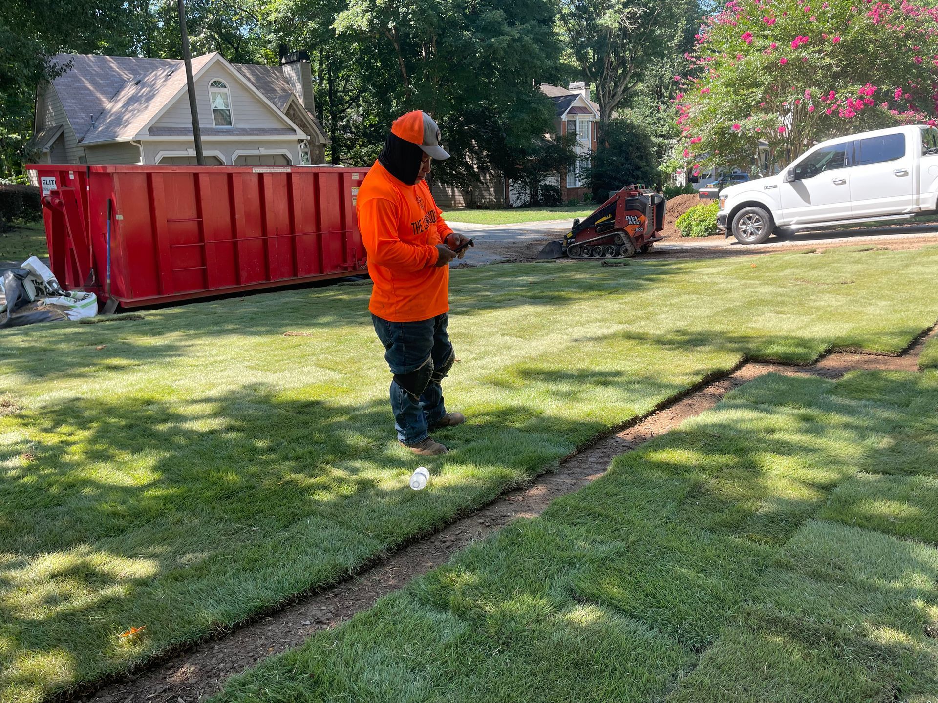 A man is standing on a lush green lawn next to a red dumpster.