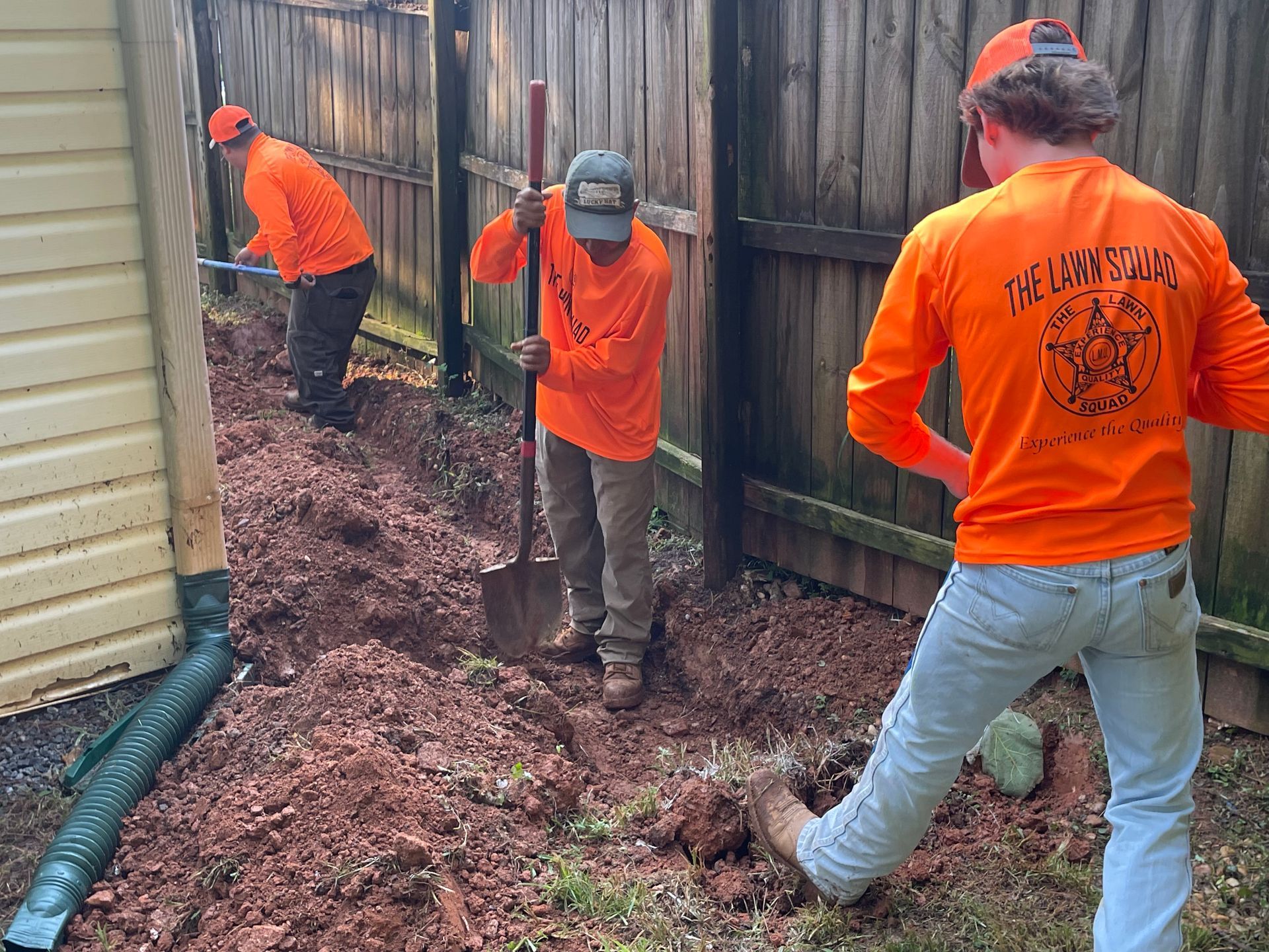 Three men in orange shirts are digging in the dirt in front of a wooden fence.