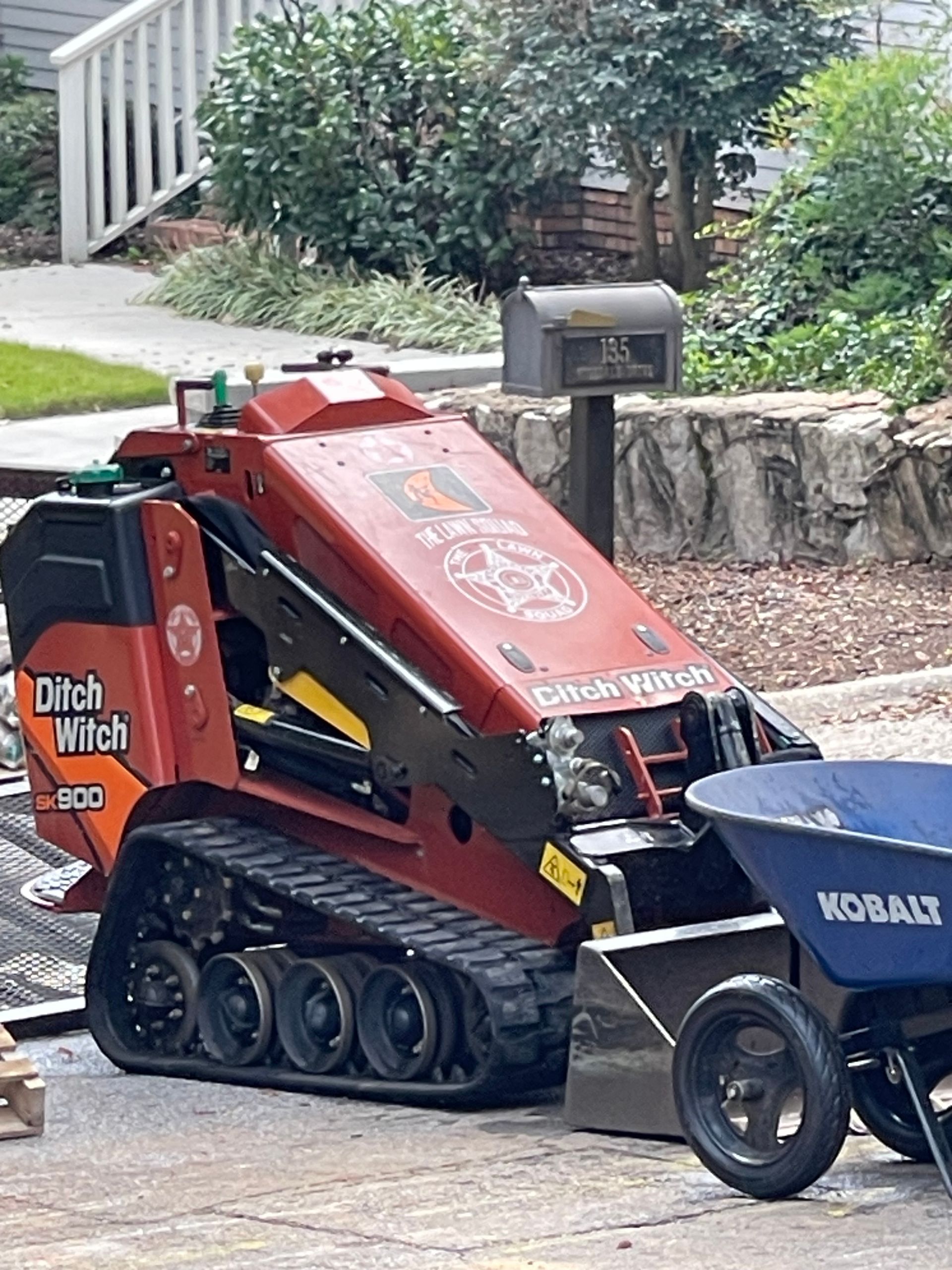 A red ditch winch is parked next to a blue wheelbarrow