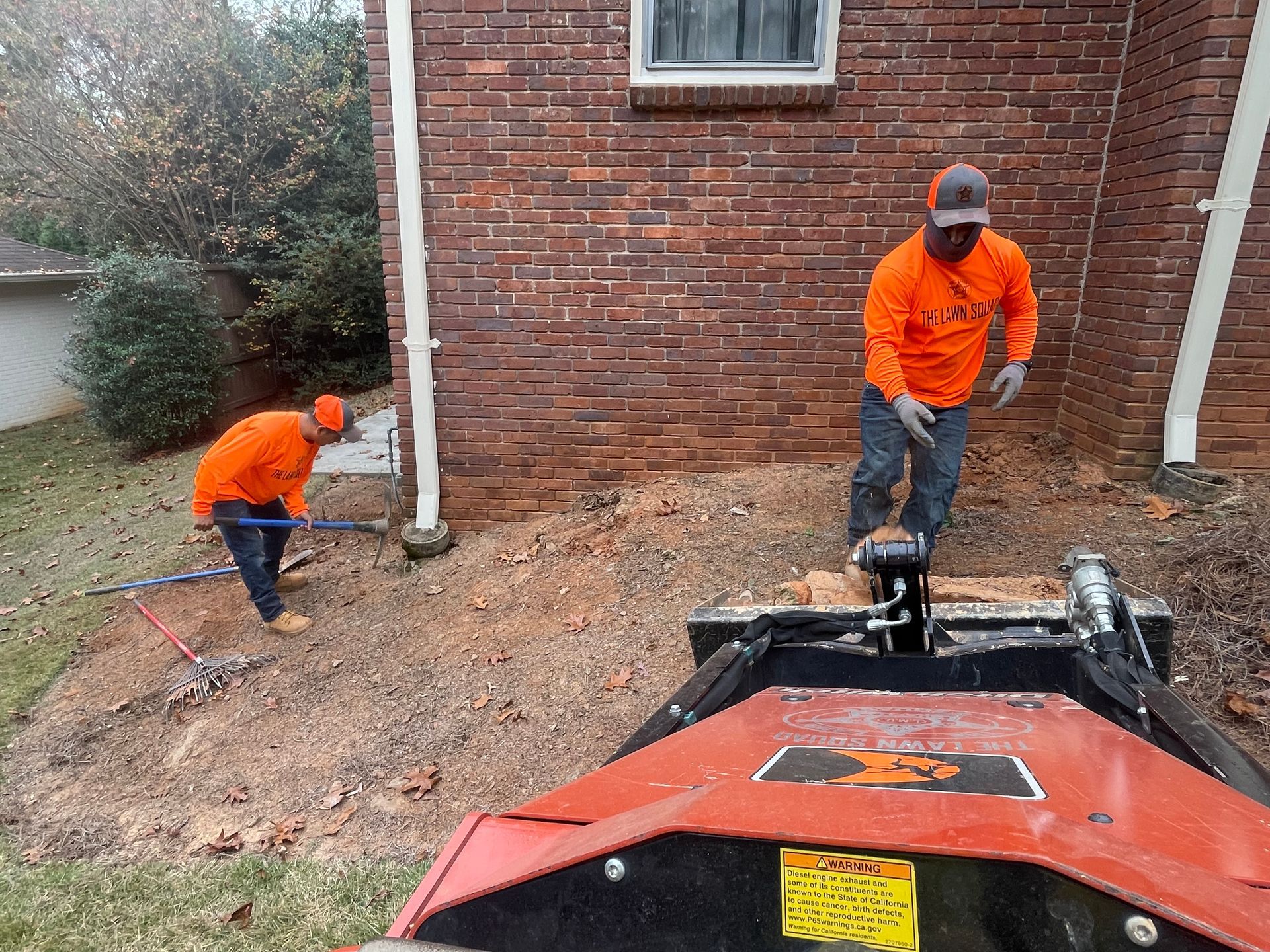 Two men are working on a tree stump in front of a brick house.